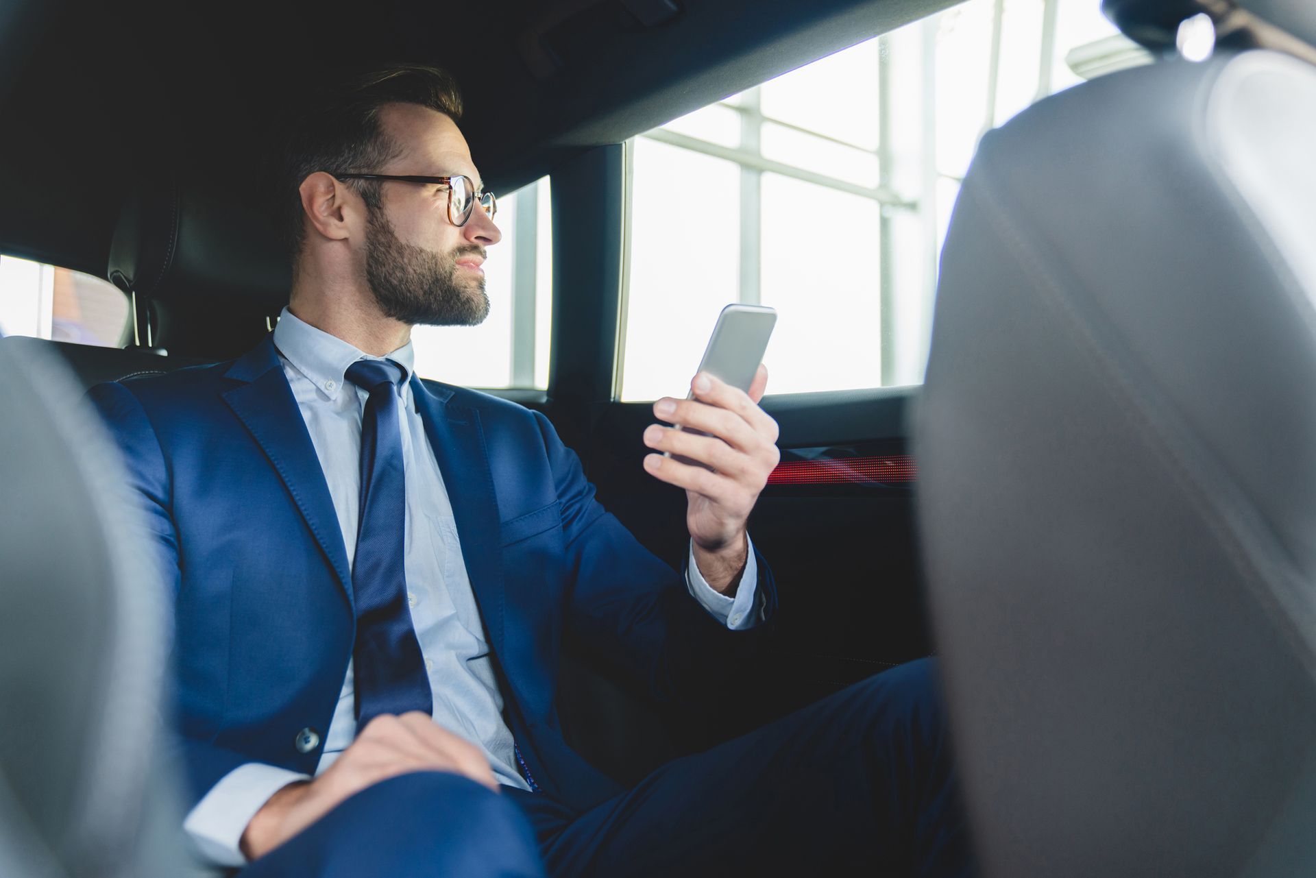 A man in a suit and tie is sitting in the back seat of a car looking at his cell phone.