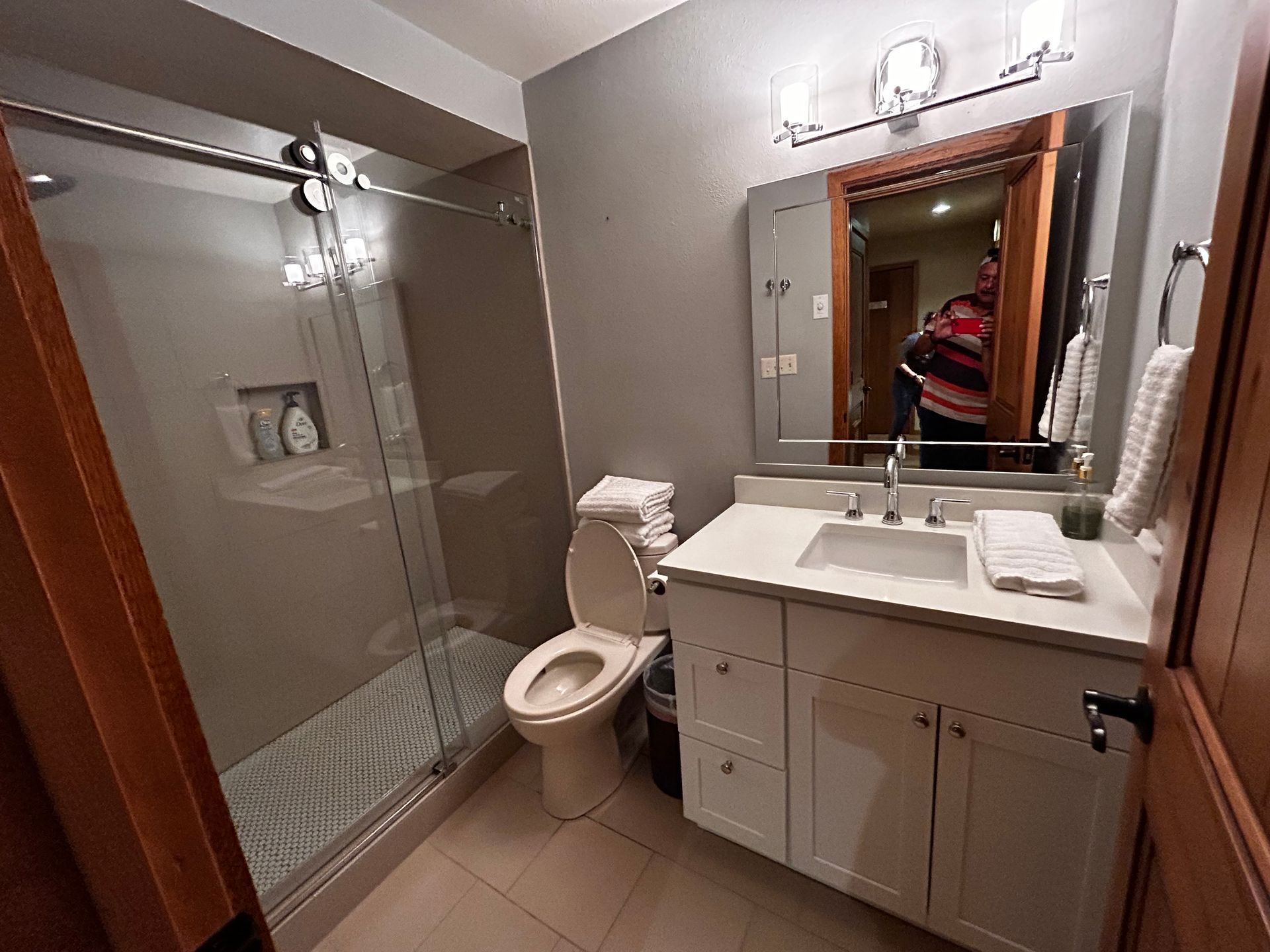 Bathroom with glass shower, toilet, white vanity, and mirror reflecting a person.
