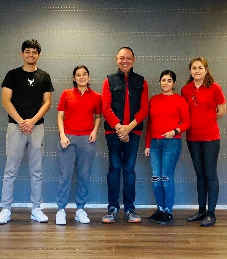 Five people stand in front of a gray wall. Three wear red shirts, one a black vest, and one a black t-shirt.