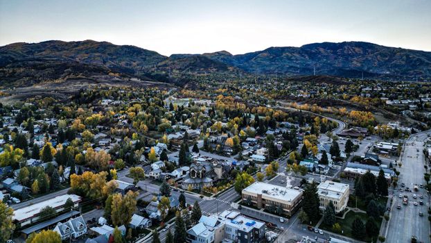 Aerial view of a town nestled in a valley with mountains in the background, trees in autumn colors, and a road.