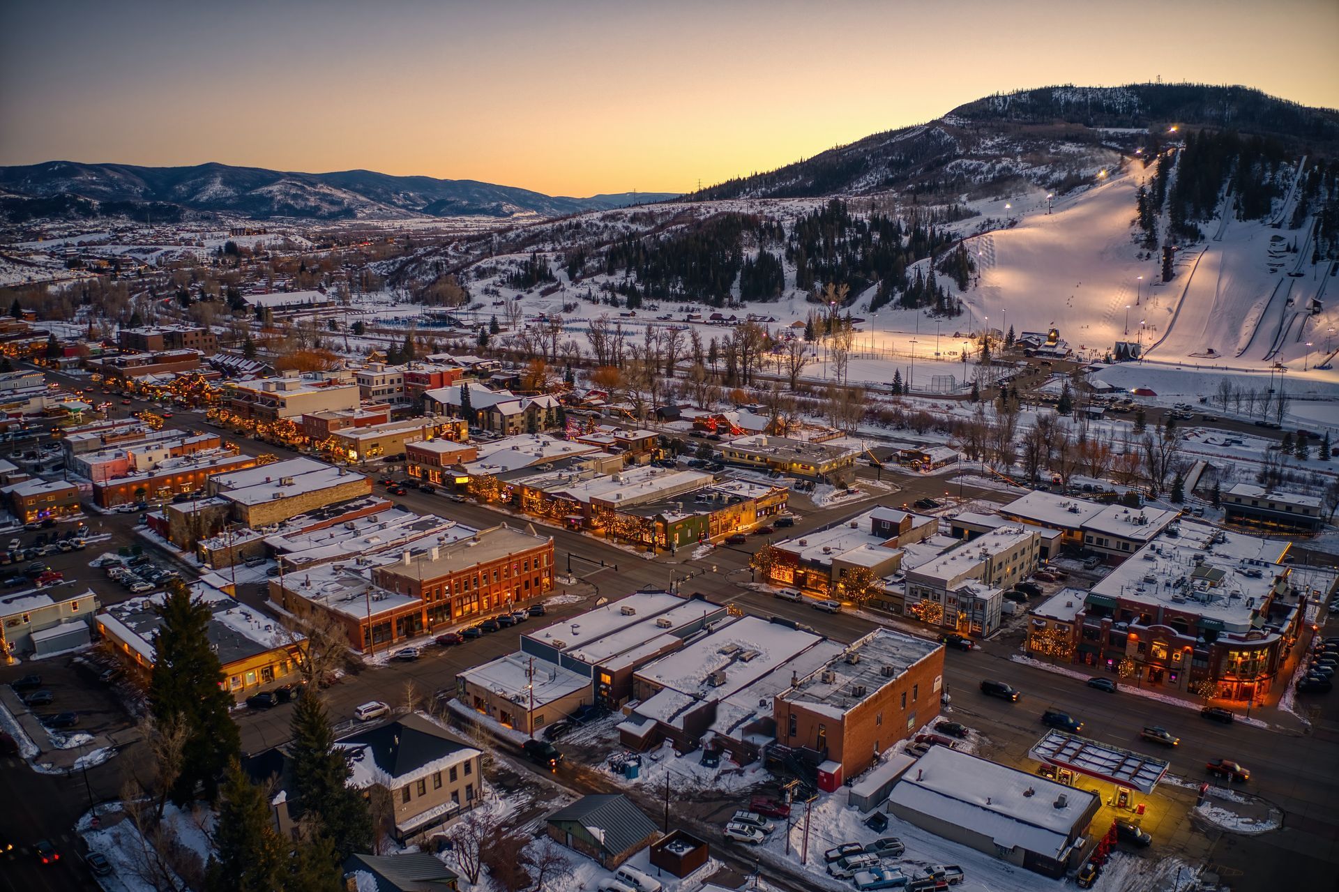 Snowy aerial view of a town at dusk. Buildings with lights, a ski slope on a hill in the background.