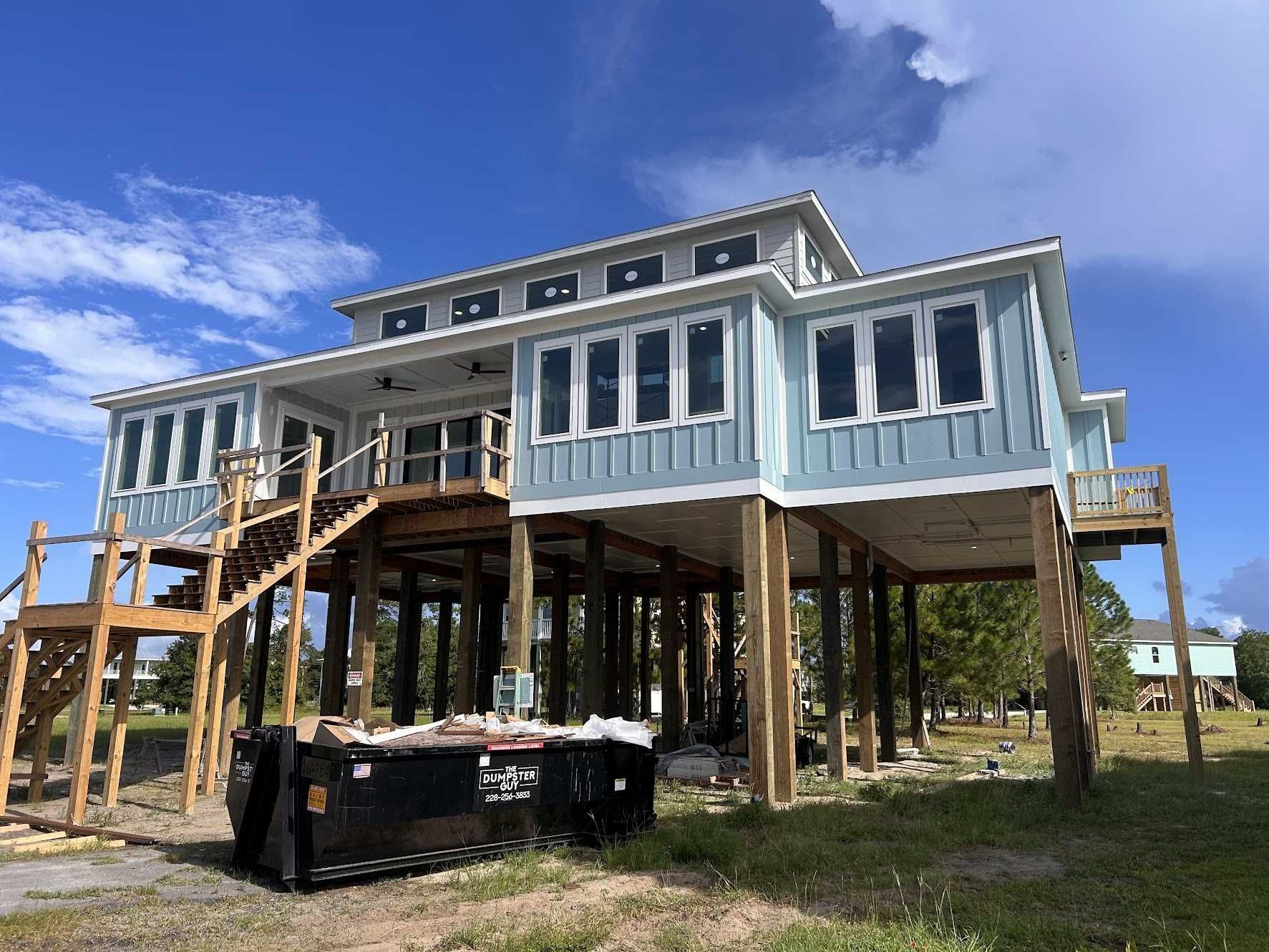 A light blue beach house on stilts under construction. Large windows, wooden stairs and supports, with a blue sky background.
