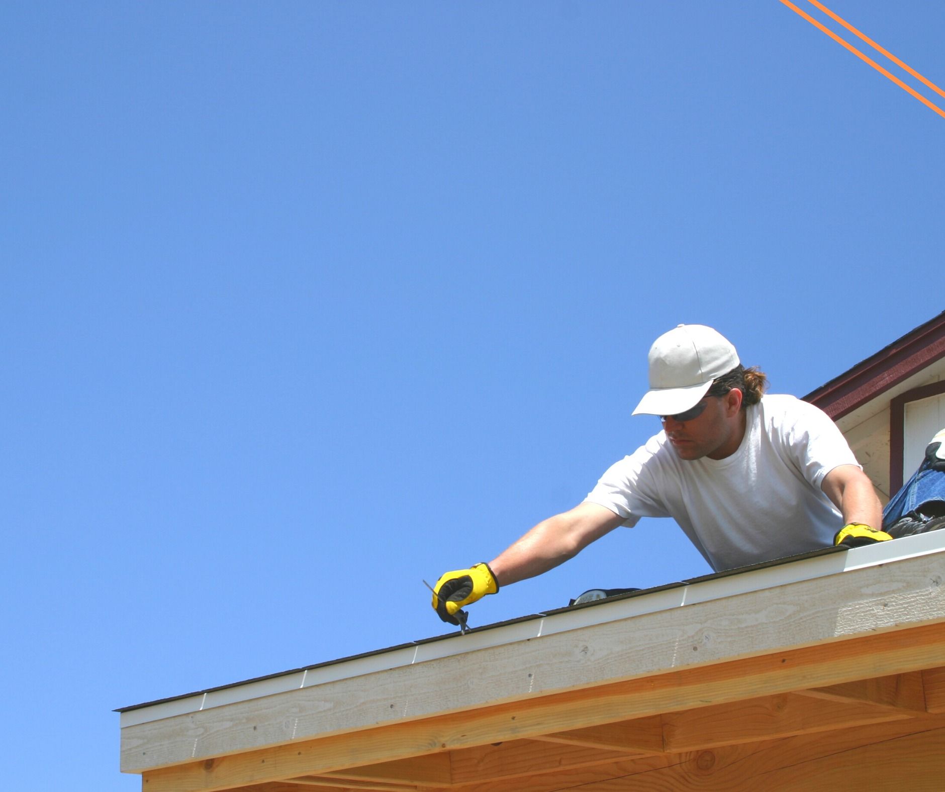 Man on a rooftop, wearing gloves and hat, leans over to work on the edge of a roof against a bright blue sky.