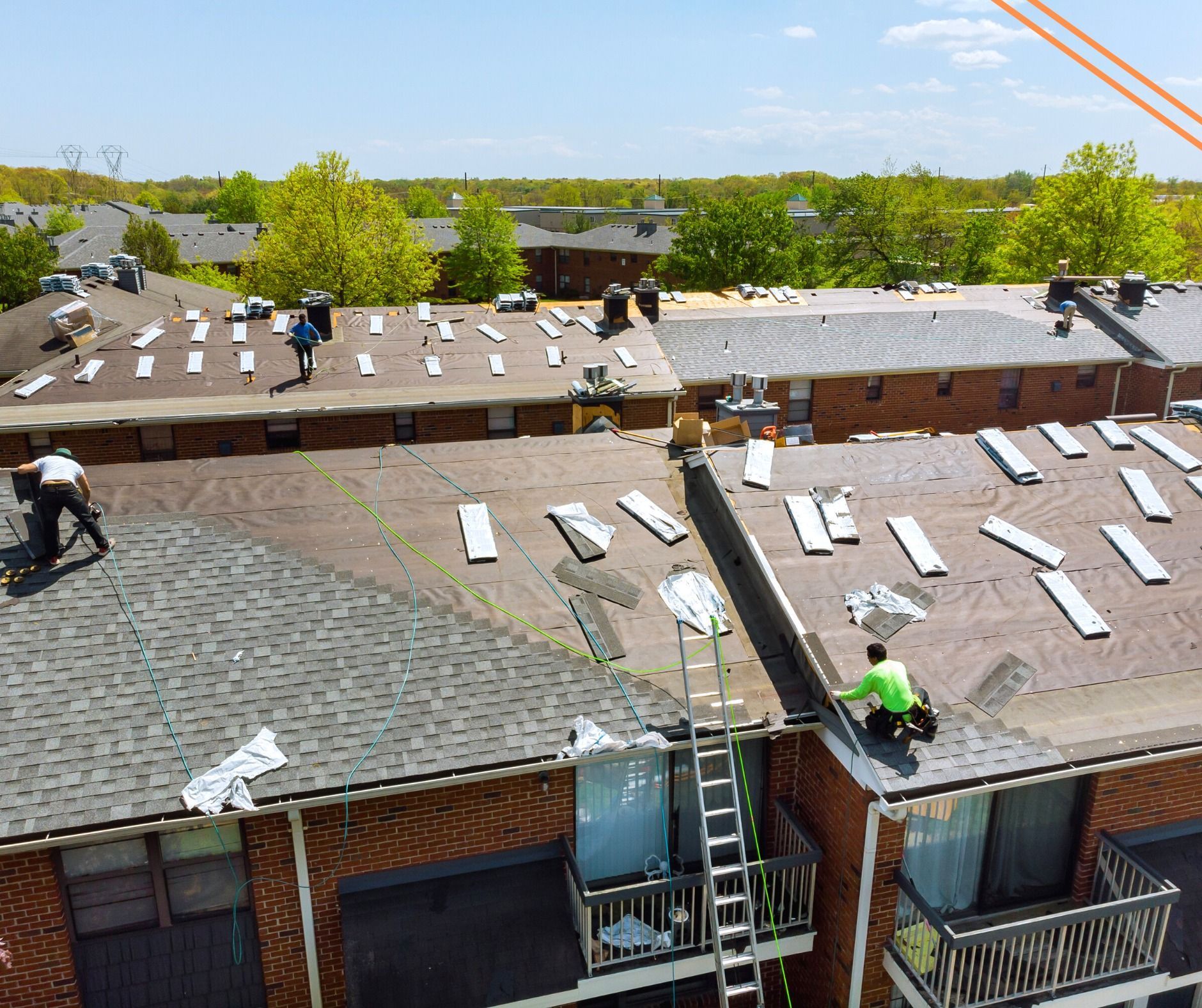 Workers replacing the roof of a brick apartment building. Several people are on the roof, with ladders and construction materials visible.