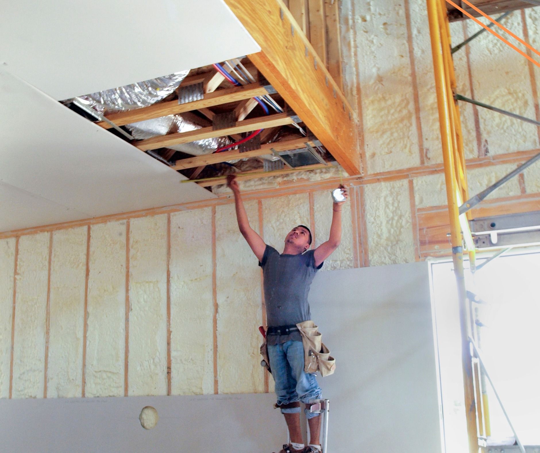 A worker on stilts installs drywall in a room under construction, reaching up toward an opening in the ceiling.