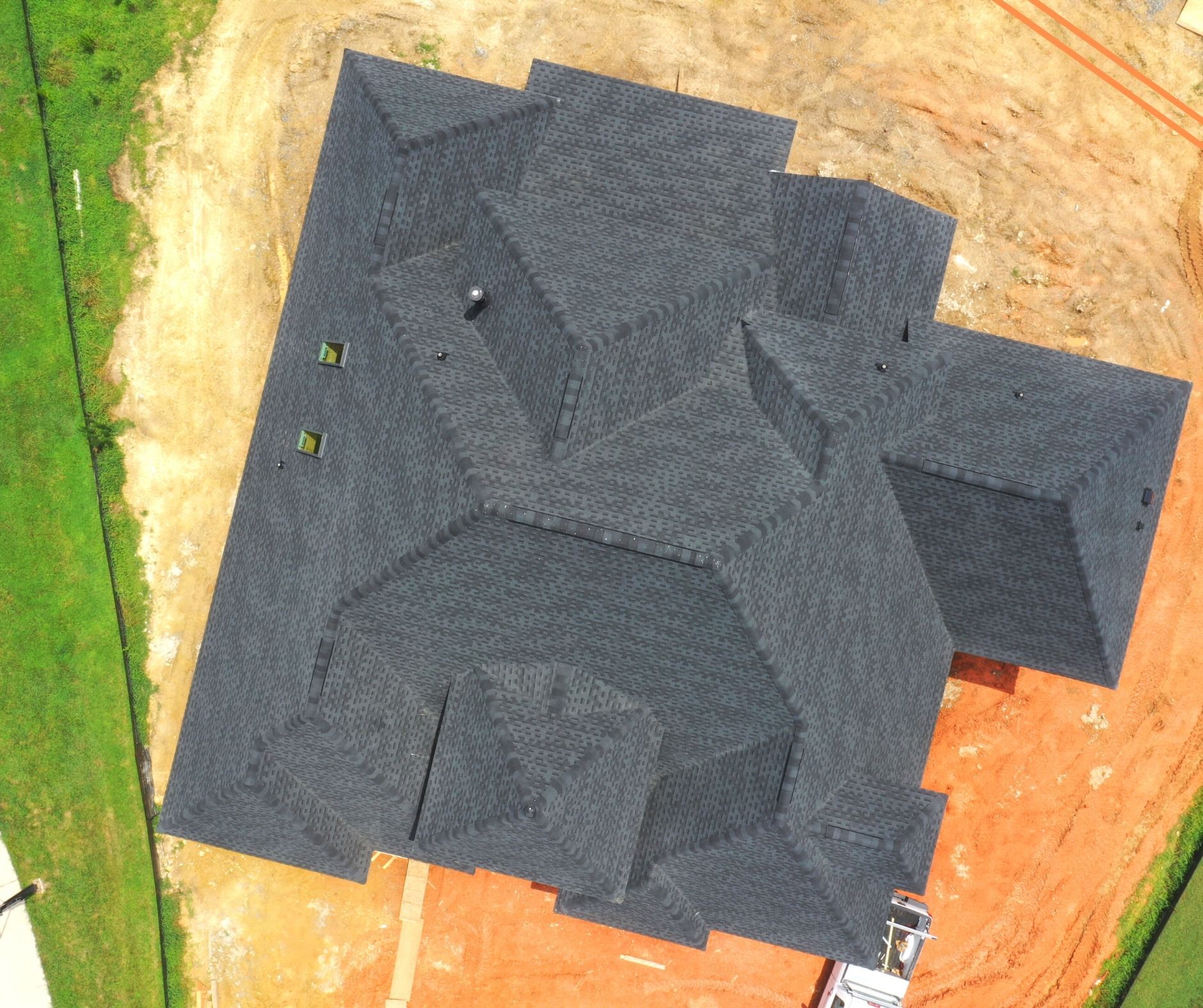 Overhead view of a house with a dark gray shingle roof. The building is on a dirt lot with grass and construction materials nearby.