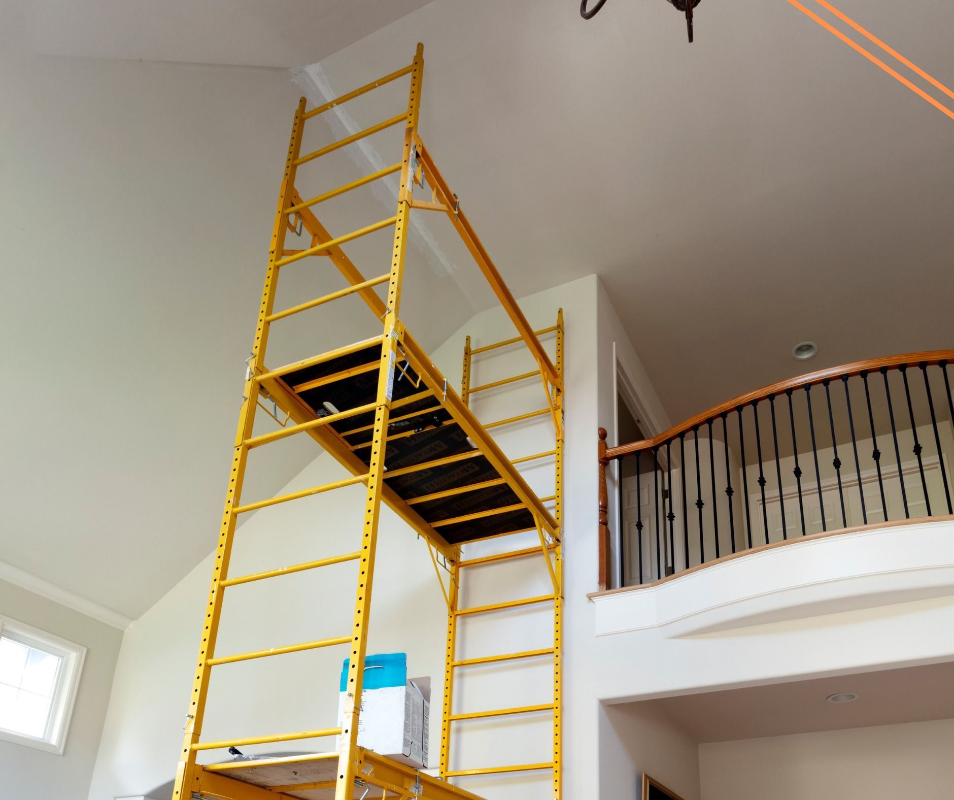 Yellow scaffolding set up inside a two-story building, near a balcony with black railings. White walls and ceiling.