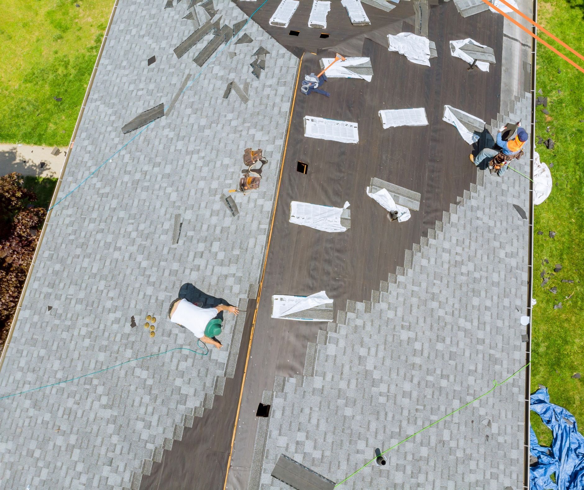 Roofers working on a gray shingle roof, removing old shingles and preparing the surface. Green lawn visible below.