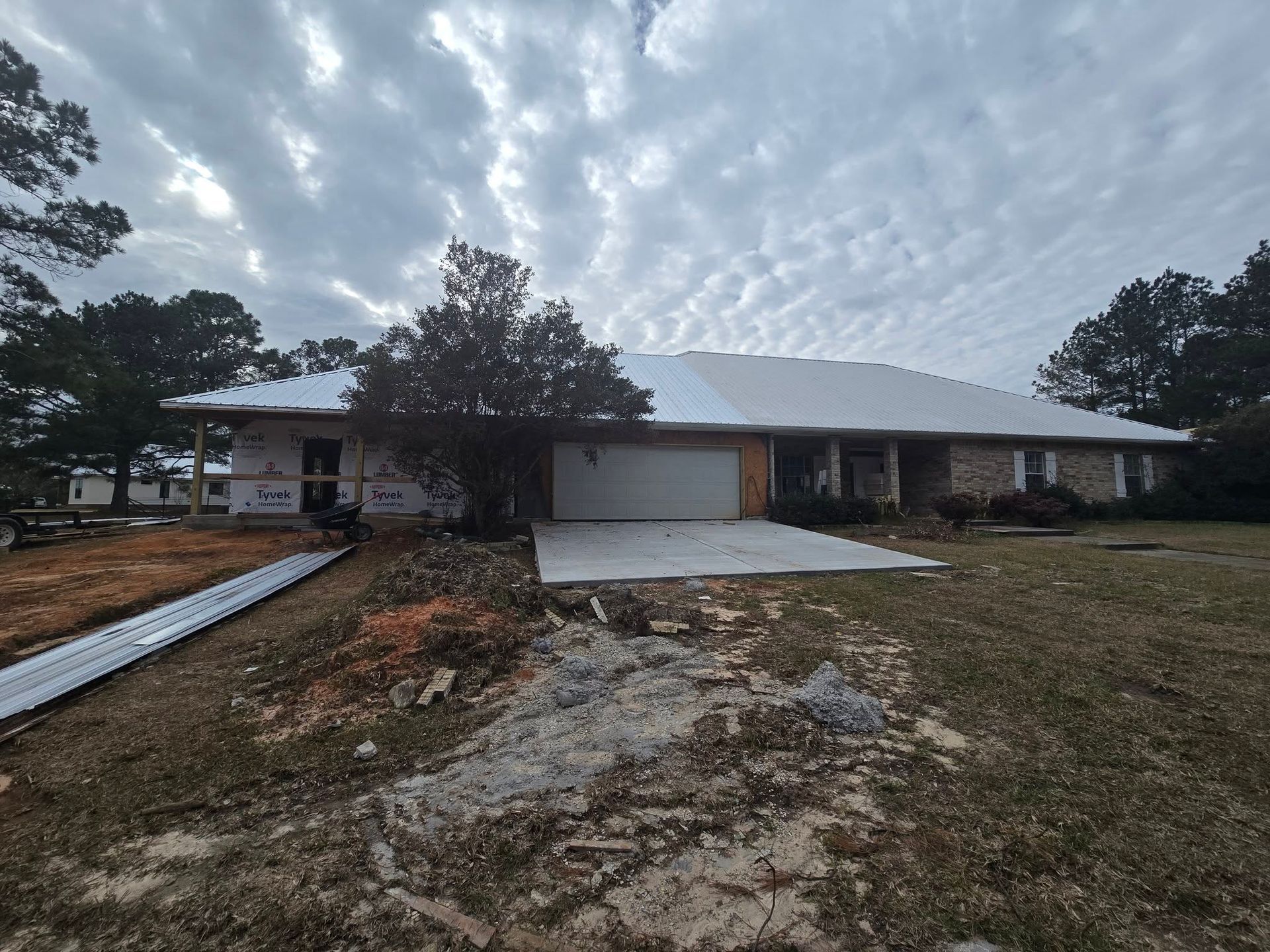 A house with a new roof being installed under a cloudy sky. The driveway is concrete, and the yard has dead grass.