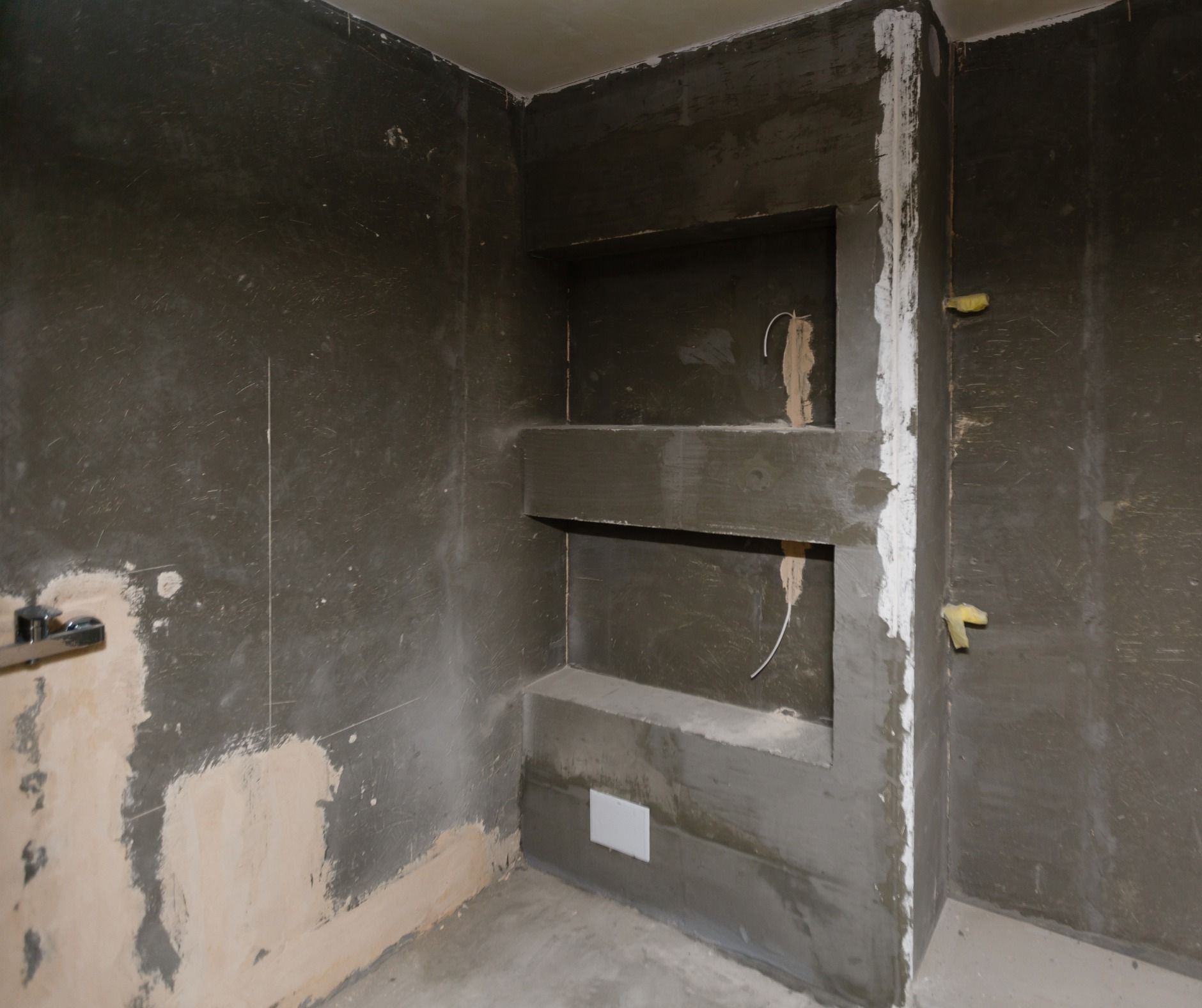 Interior of a bathroom under construction; gray walls with built-in shelves. Rough cement finish, exposed wiring.