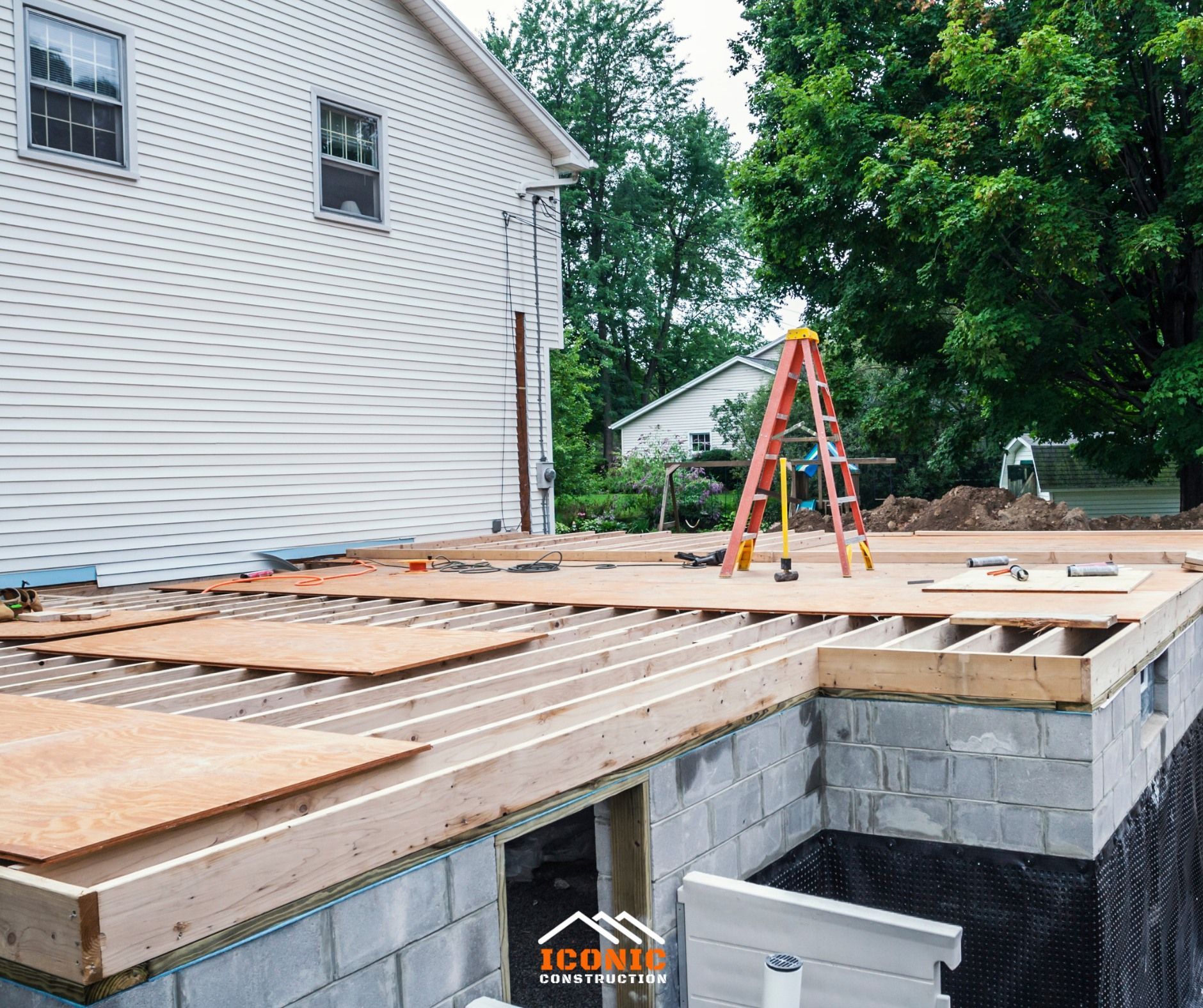 Construction site: a house with a partially built deck, wood framing, and cinder block foundation. A ladder and tools are present.