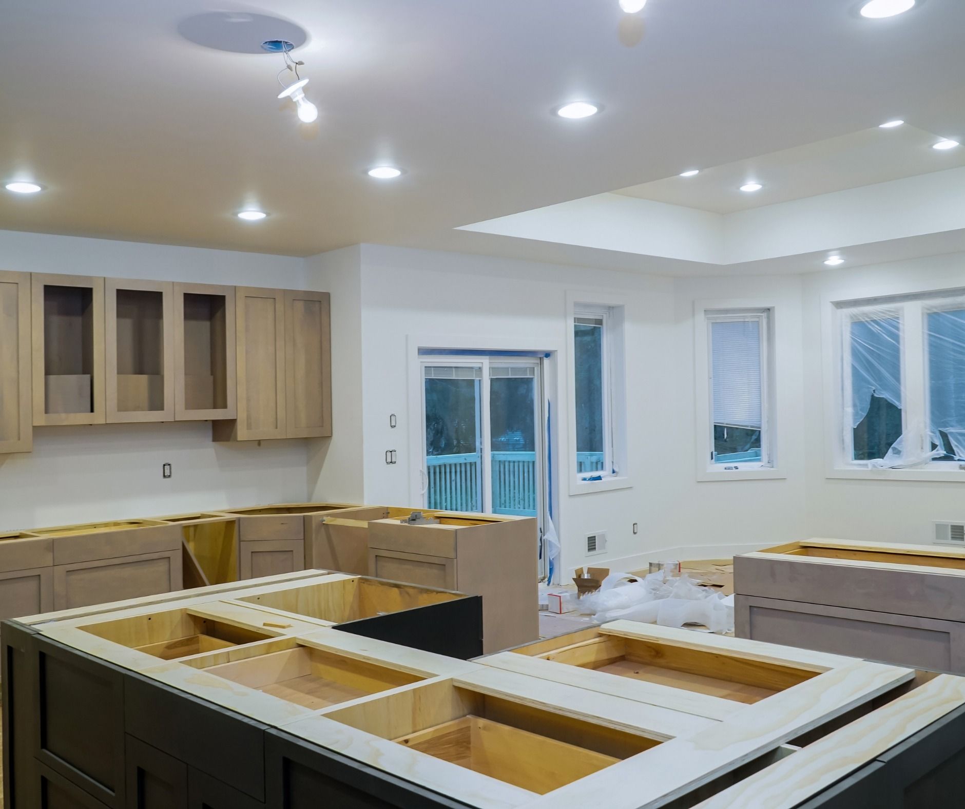 Kitchen under construction, showing unfinished cabinets, island, and recessed lighting. The room is light-colored with windows and a sliding glass door.