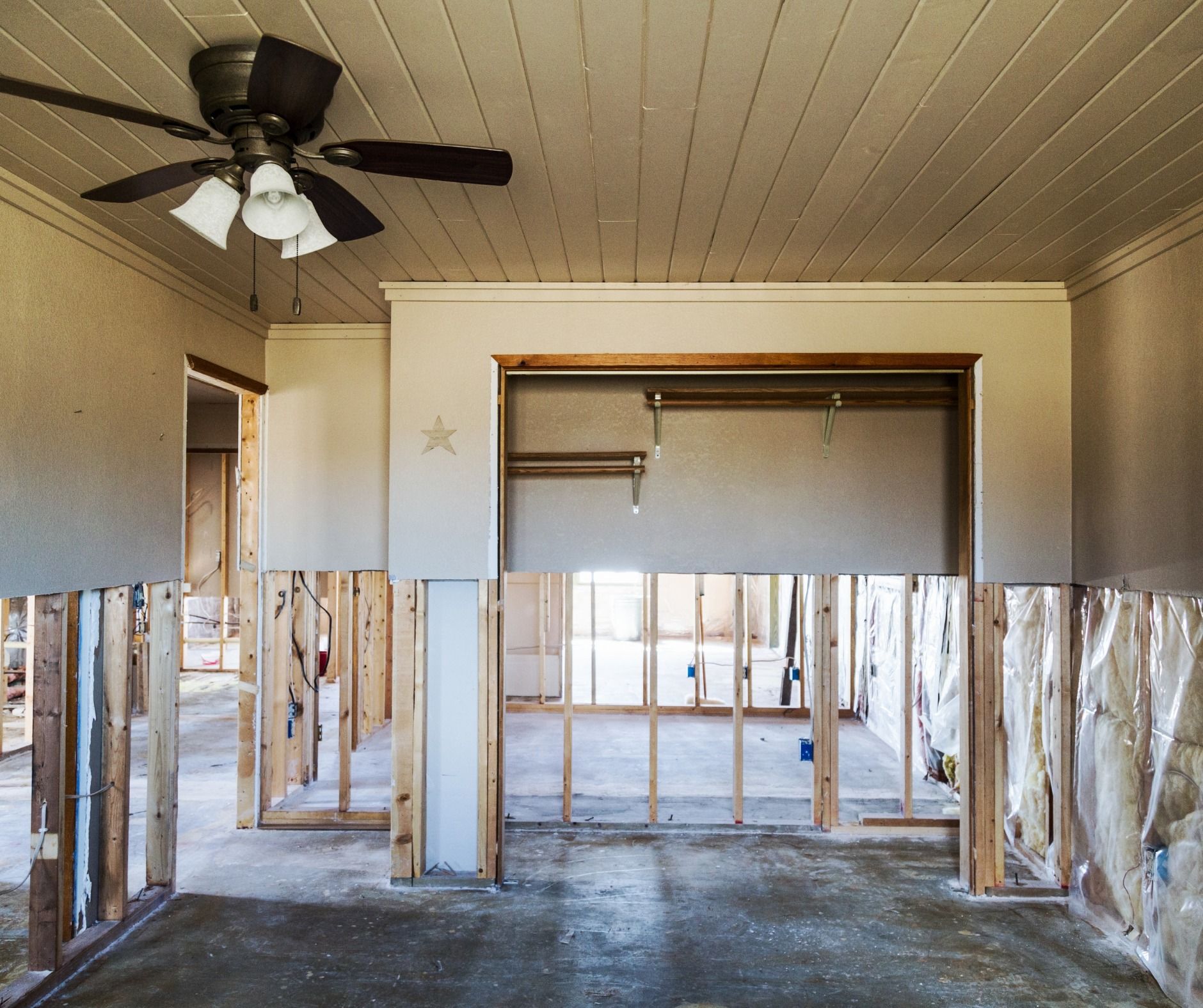 Interior of a room undergoing renovation, with exposed wall framing, ceiling fan, and open doorways.