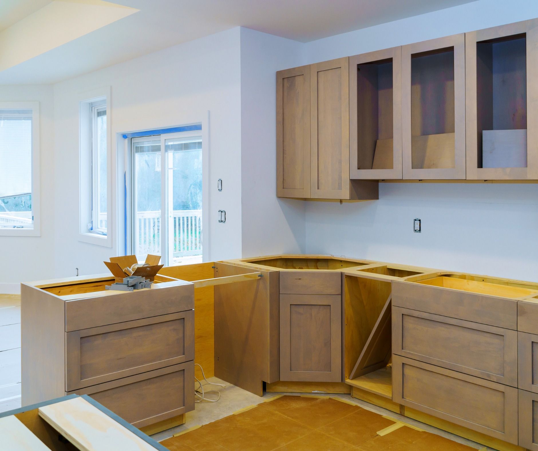 Kitchen under construction with unfinished light brown cabinets and upper cabinets with glass doors; light and airy setting.