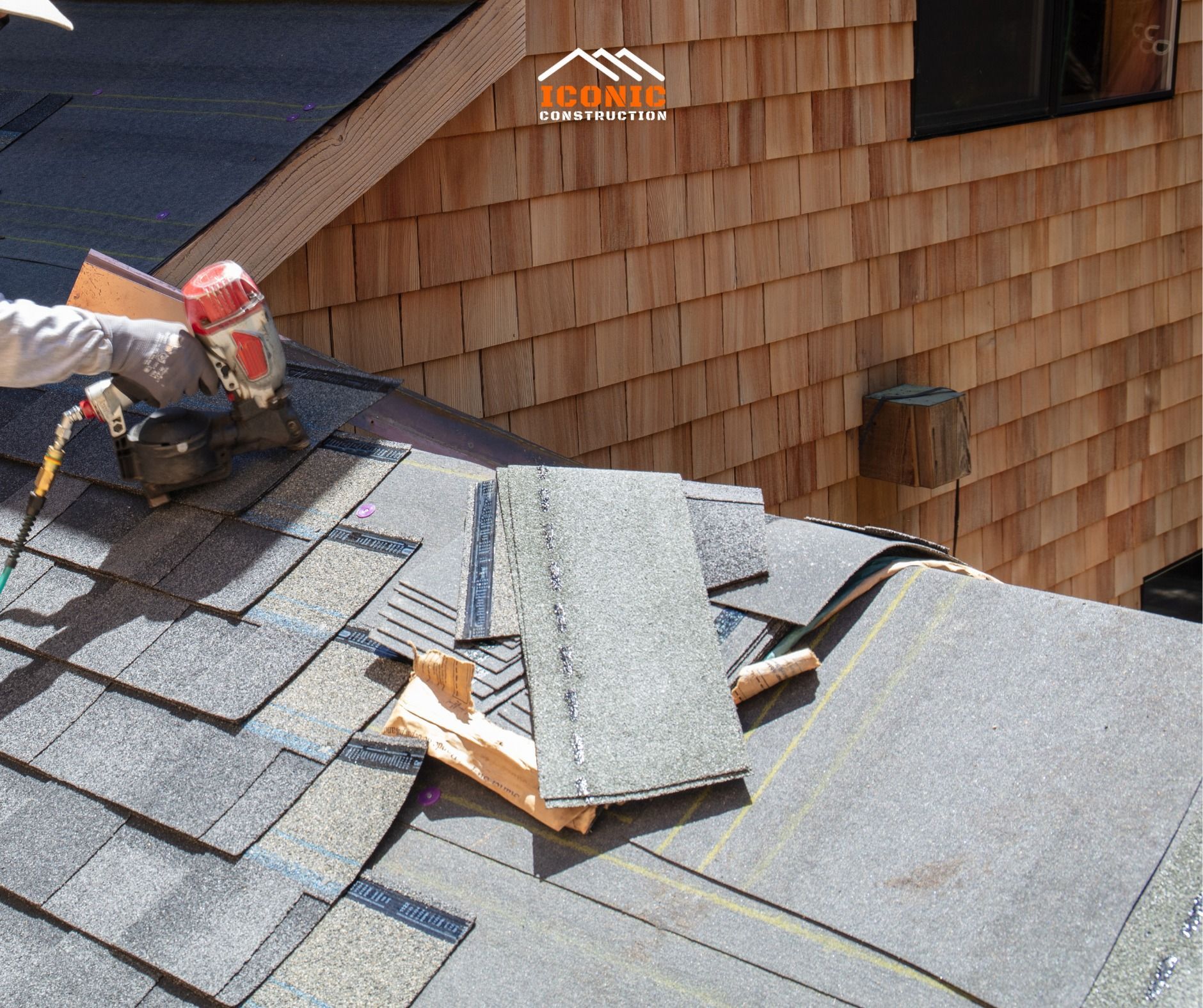 Roofer using a nail gun on a gray shingle roof with a cedar shake wall in the background, construction scene.