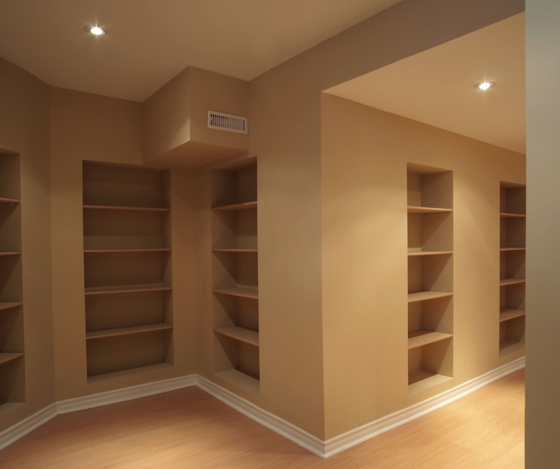 Empty room with built-in tan bookshelves along two walls and a corner, lit by recessed ceiling lights.