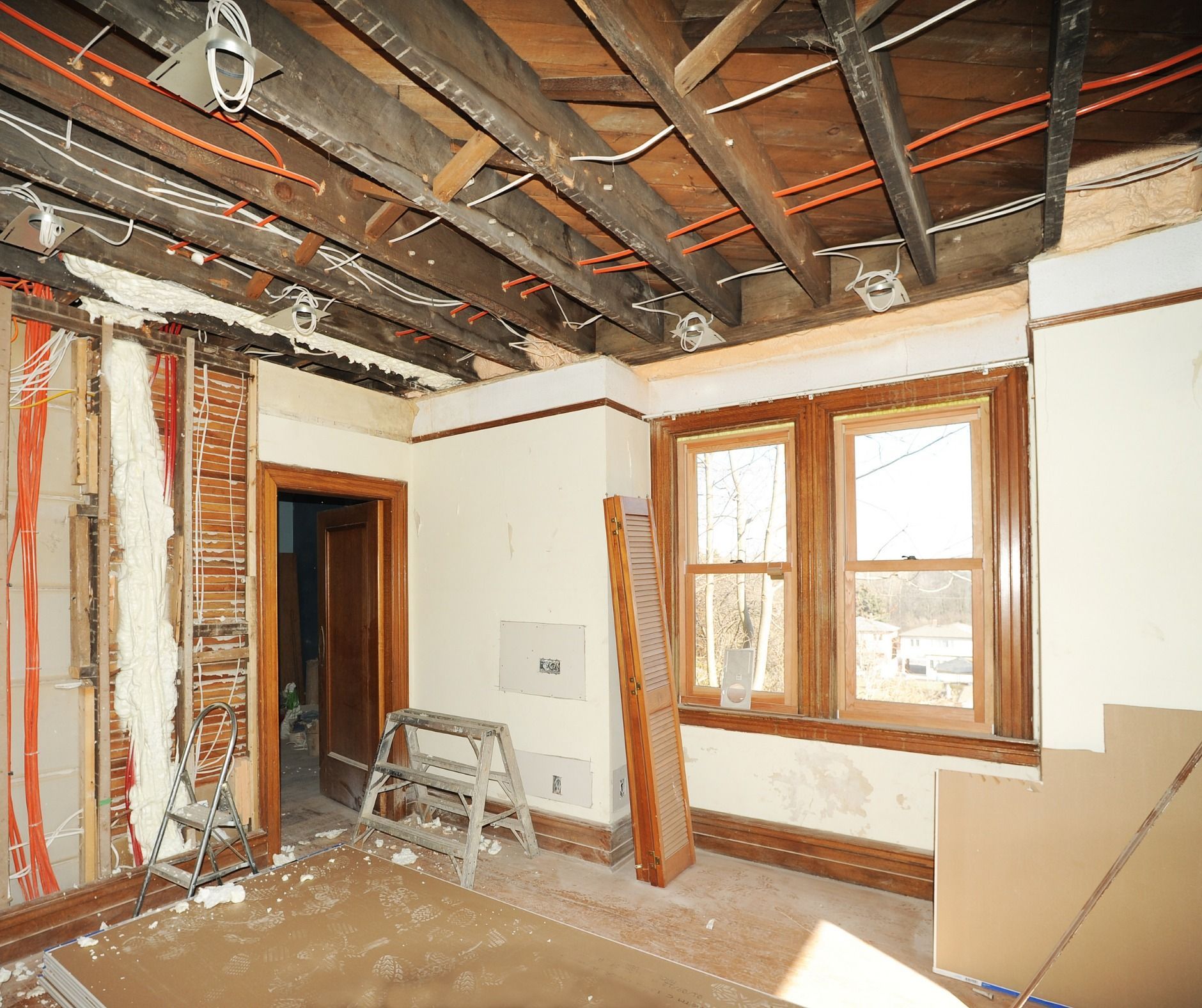 Room under renovation: exposed ceiling beams, damaged walls, ladder, and windows with wood trim.