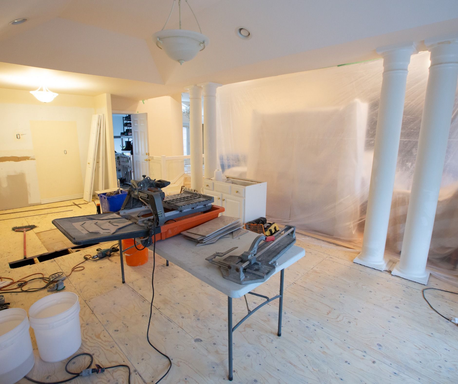 Interior of a room under renovation. Tools, a table, and plastic sheeting are present. White columns and light fixtures are visible.