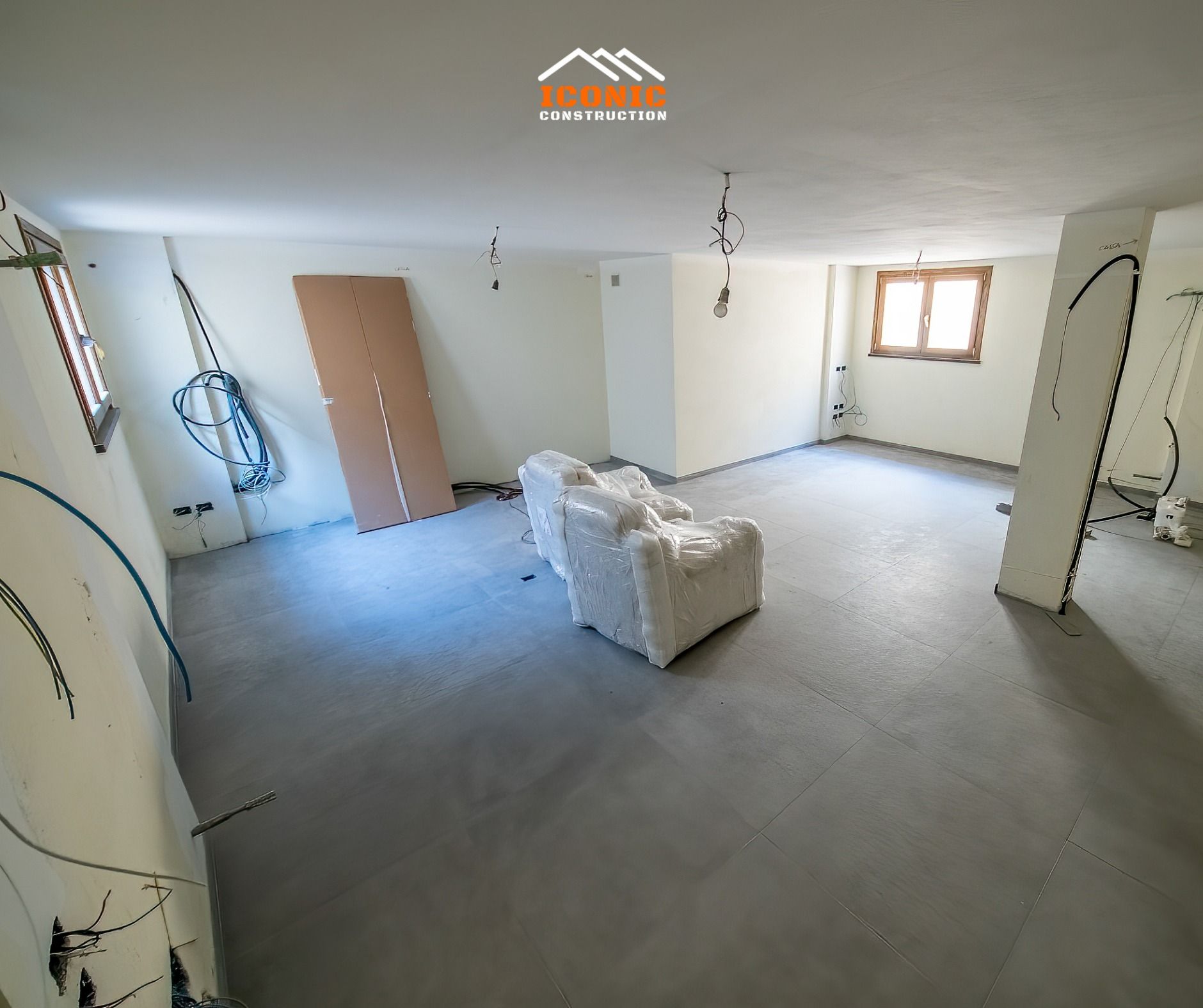 A room under construction with exposed wiring and a sofa covered in white packing material. Gray floor, white walls, small windows.