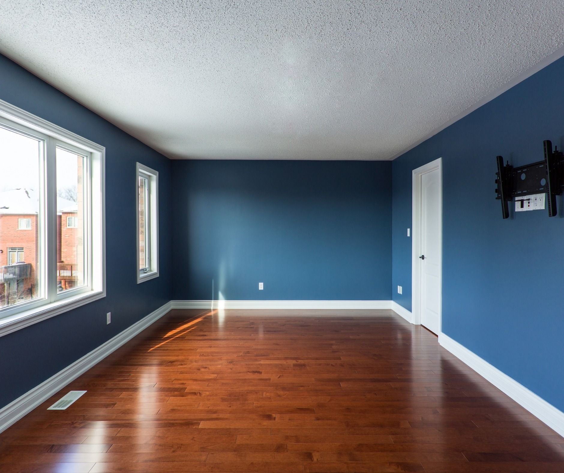 Empty room with hardwood floors, blue walls, white trim and door, and windows. A TV wall mount is visible on one wall.