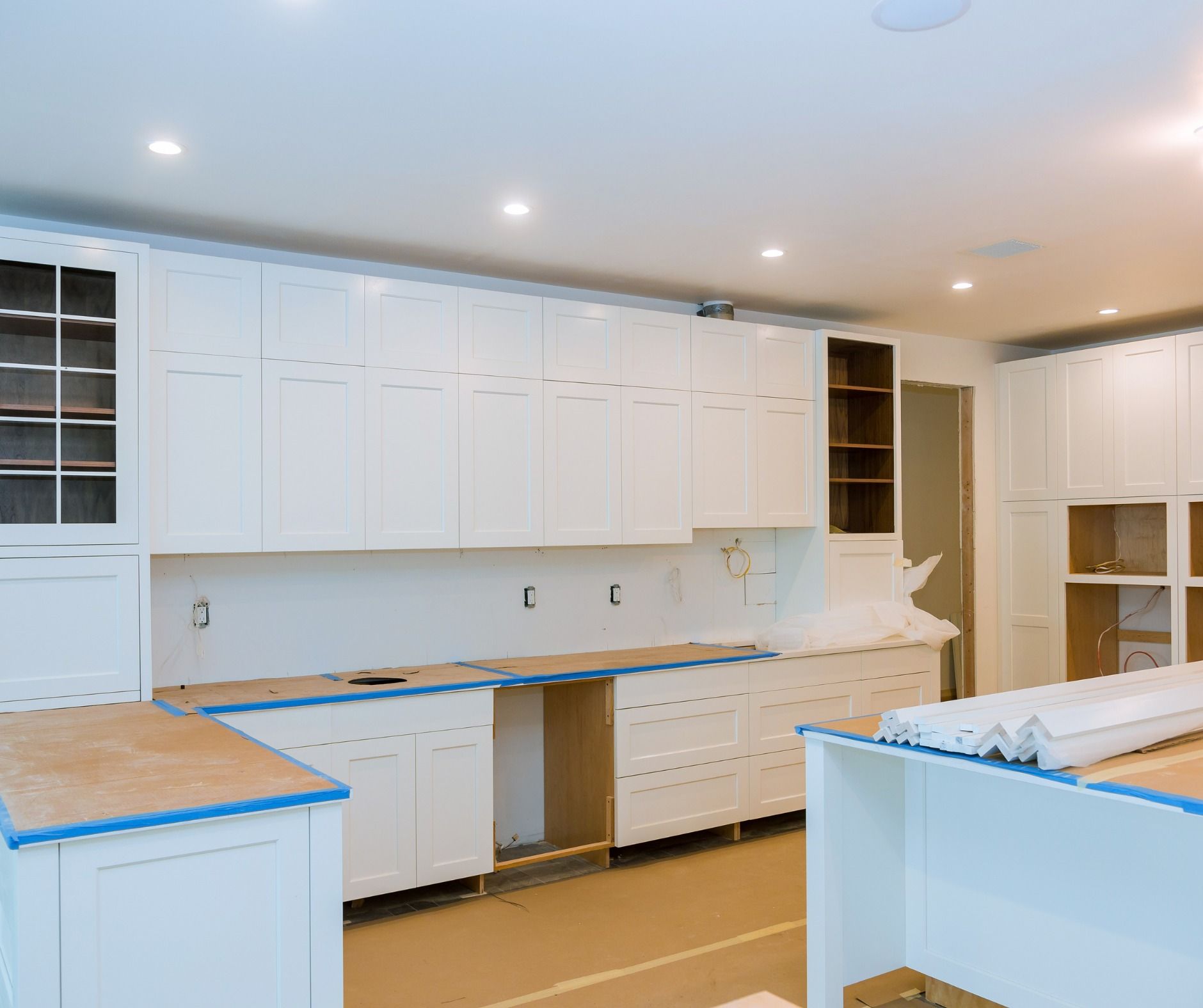 White kitchen cabinets being installed in a room with wooden flooring. Some cabinets are open and unfinished.
