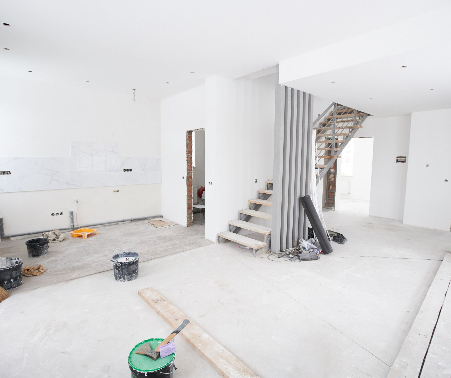Interior under renovation, with bare walls, concrete floors, exposed staircase, and construction materials.