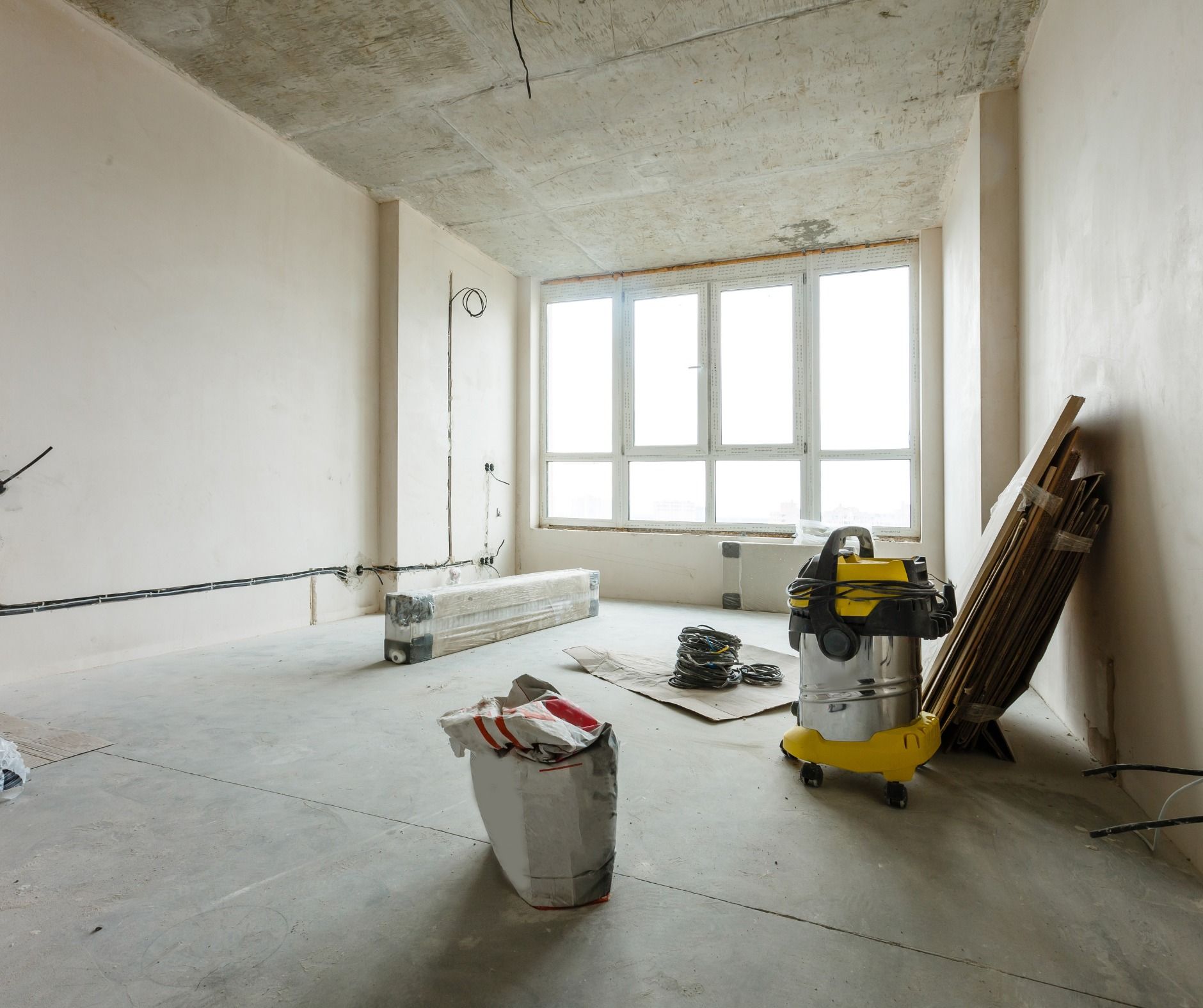 Room under construction with concrete floors and walls, a large window, and construction materials like a vacuum cleaner and wood.