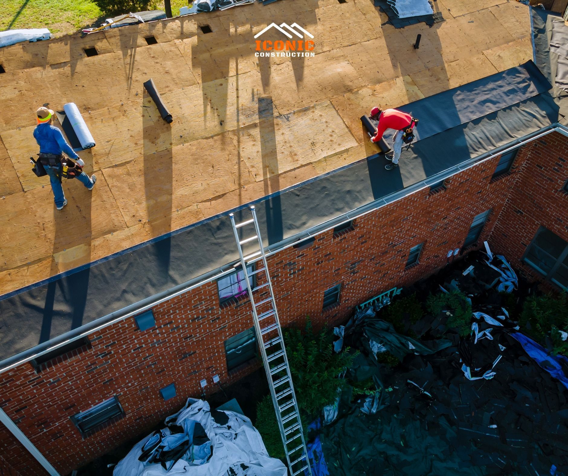 Two roofers installing roofing material on a brick building, with a ladder and rolls of material visible.