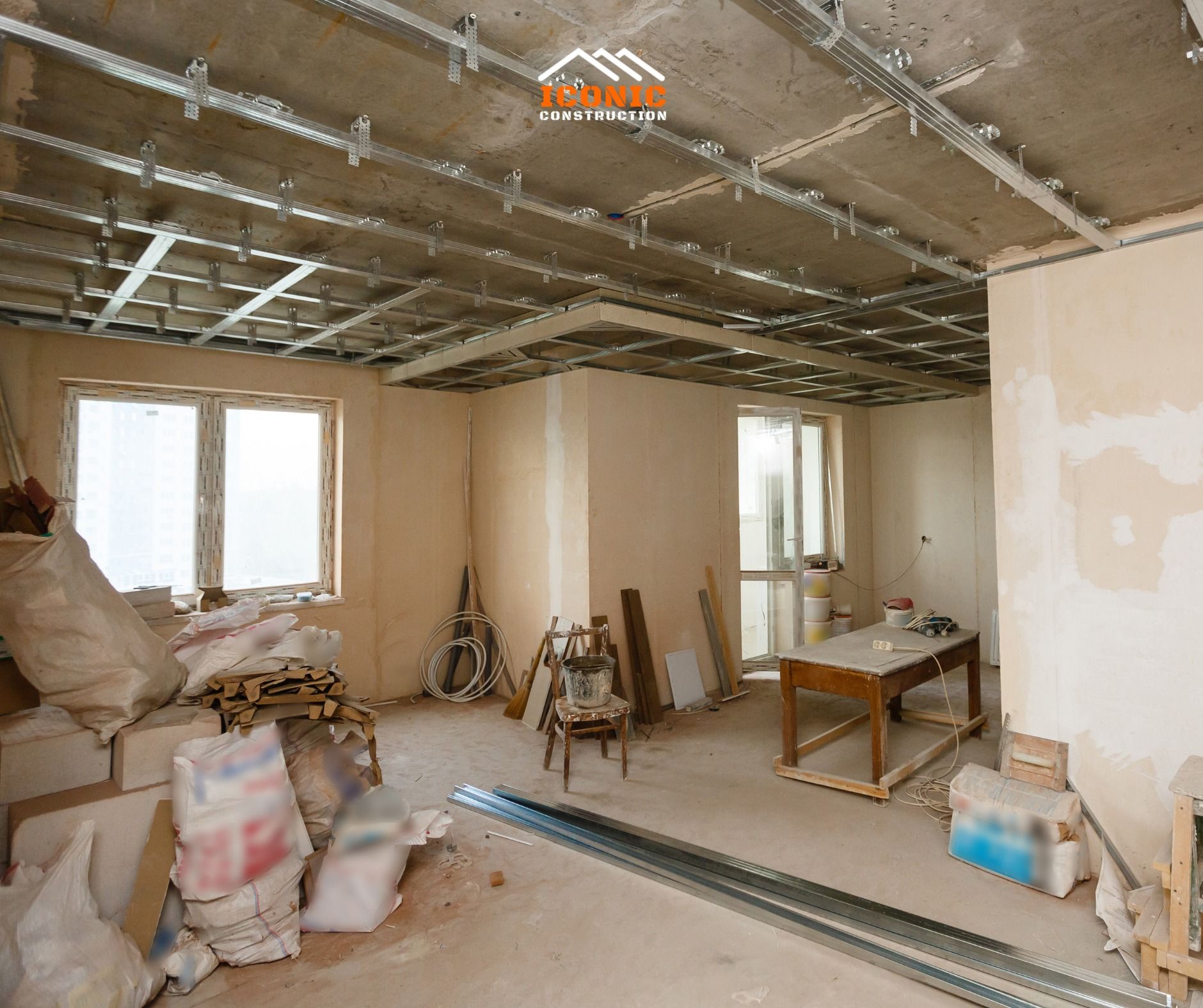 Interior of a room under construction, showing exposed metal ceiling grid, windows, and scattered building materials.