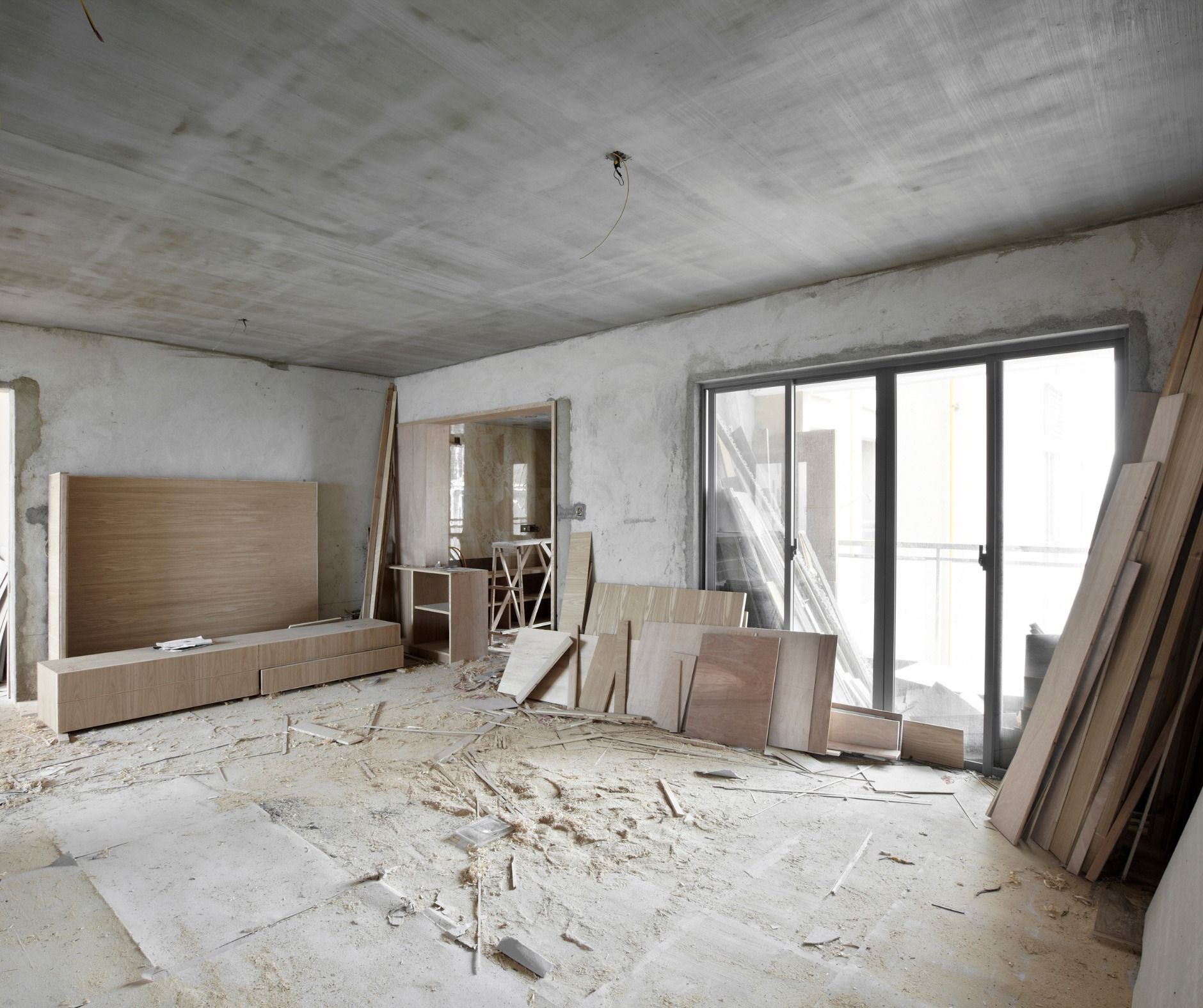 Interior room under construction with unfinished walls, flooring, and materials scattered throughout. A sliding glass door and doorway are visible.