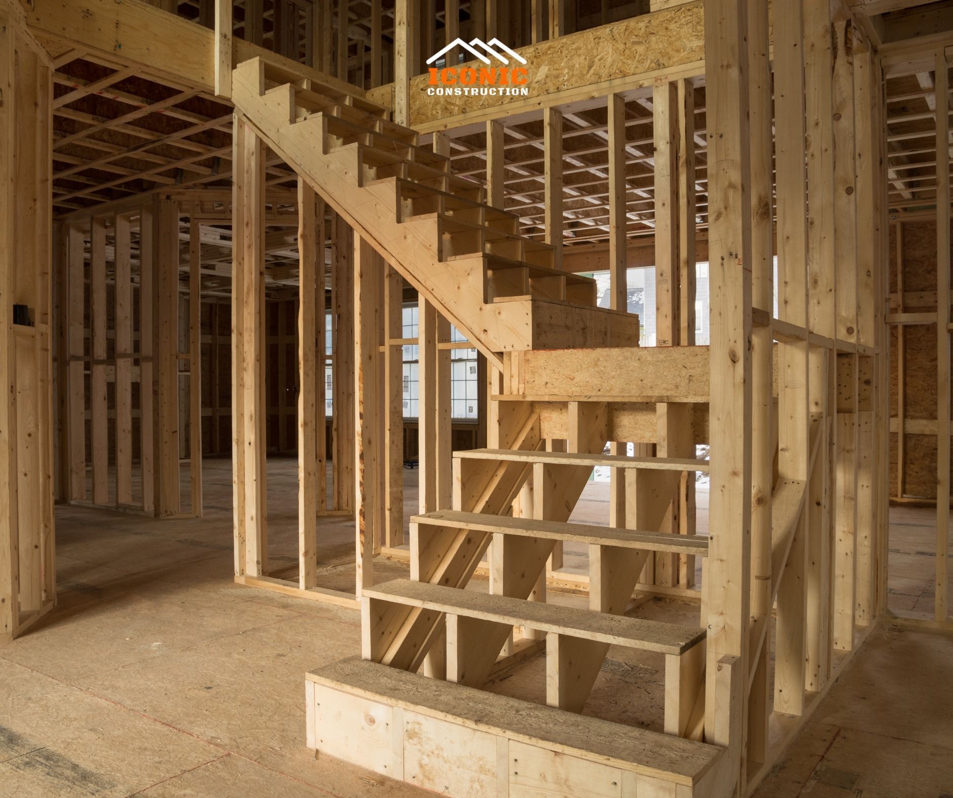 Interior view of a home under construction, showing a wooden staircase framed with exposed beams and studs. The unfinished floor is visible.