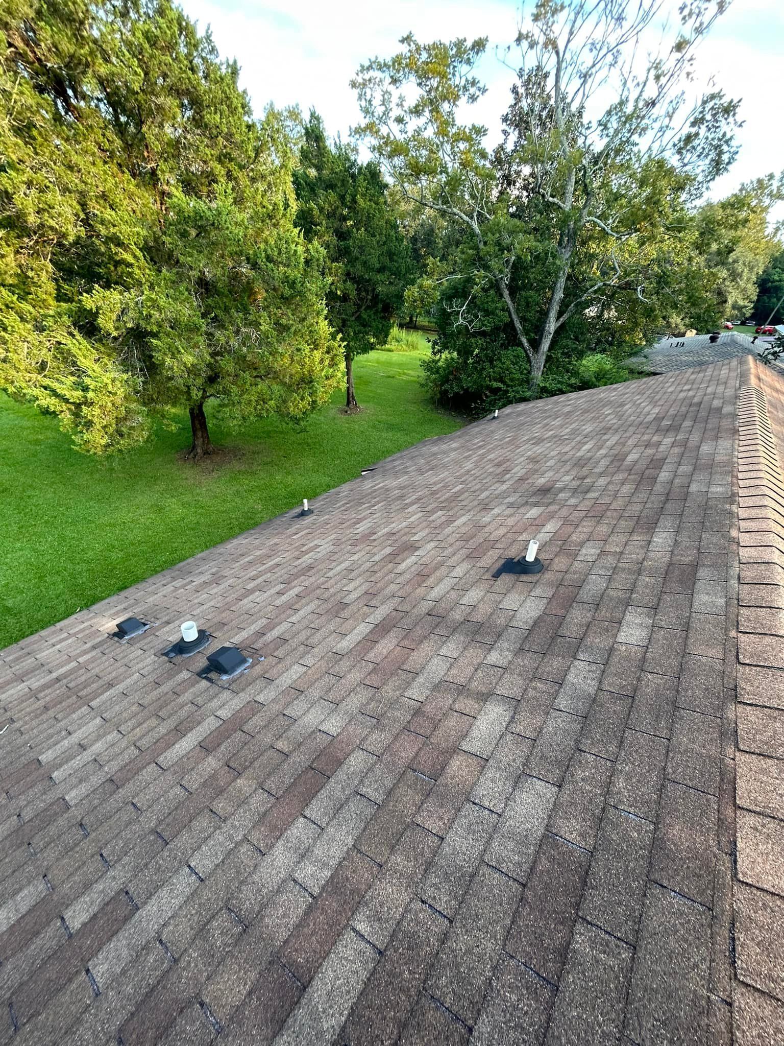 A rooftop covered in brown shingles, surrounded by green trees and grass on a sunny day.
