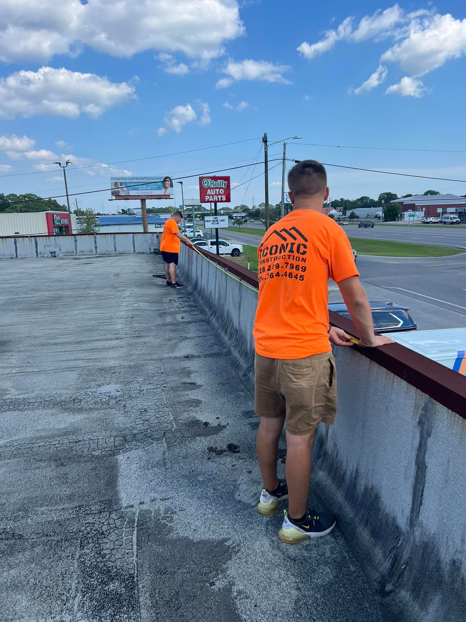 Two young men in orange shirts and shorts stand on a rooftop, looking out at a commercial area on a sunny day.