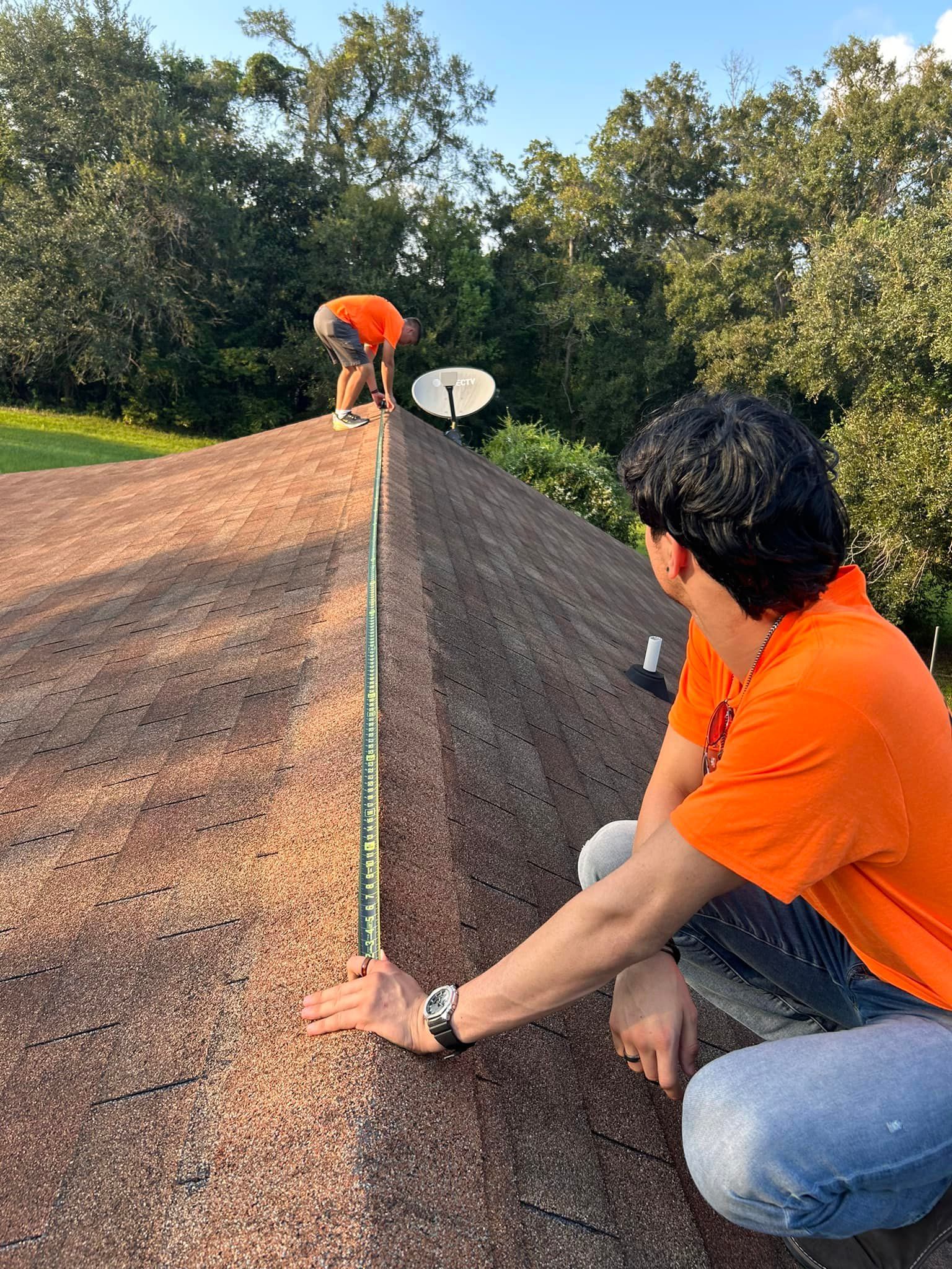 Two roofers in orange shirts assess and measure a brown shingled roof; one kneels, another stands on the roof peak.