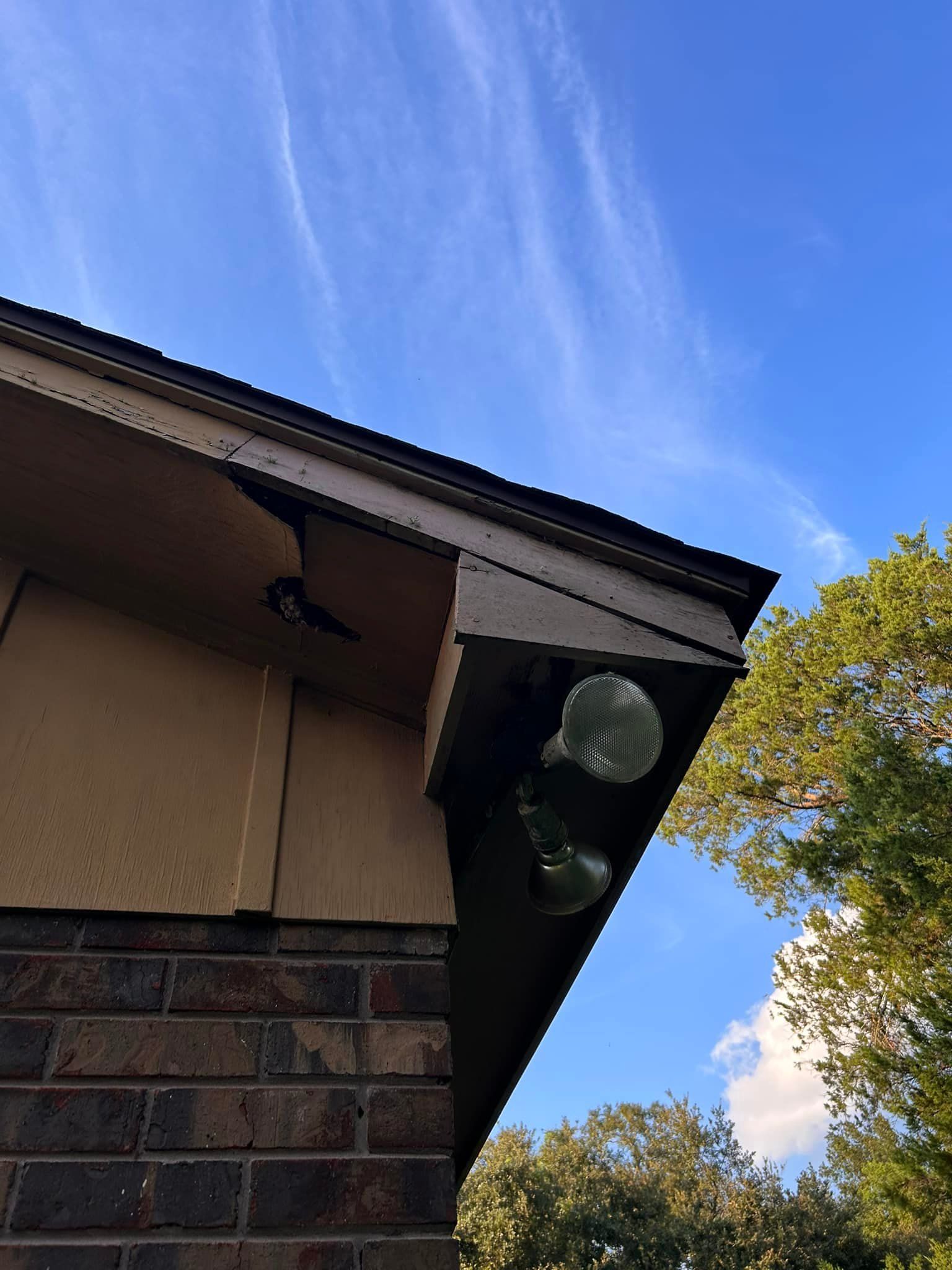 Corner of a house with a brown roof overhang, tan siding, and brick below. Two green glass globe lights are mounted. Blue sky.