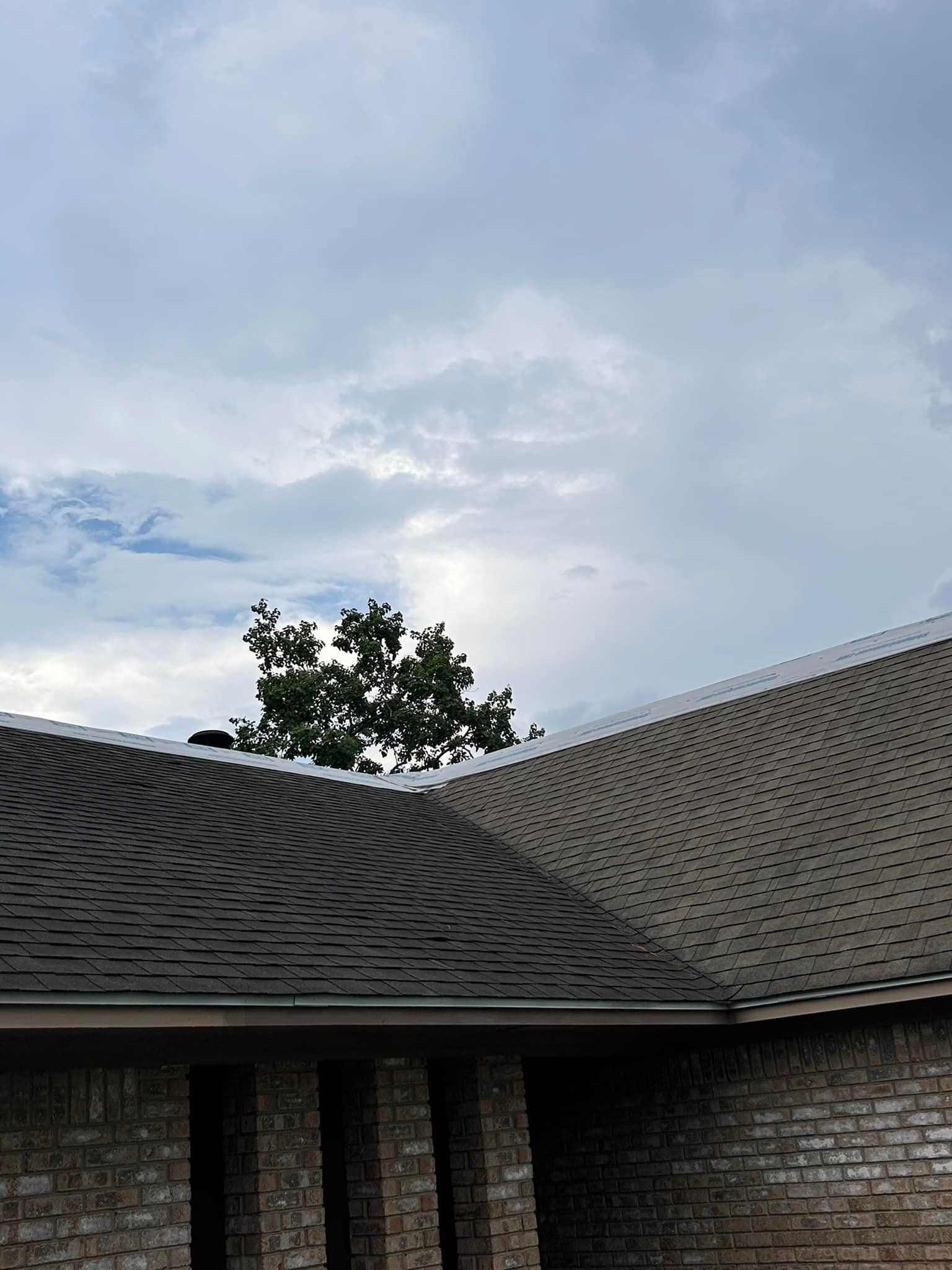 Roofline of a brick house with a tree peeking over it, against a cloudy sky.