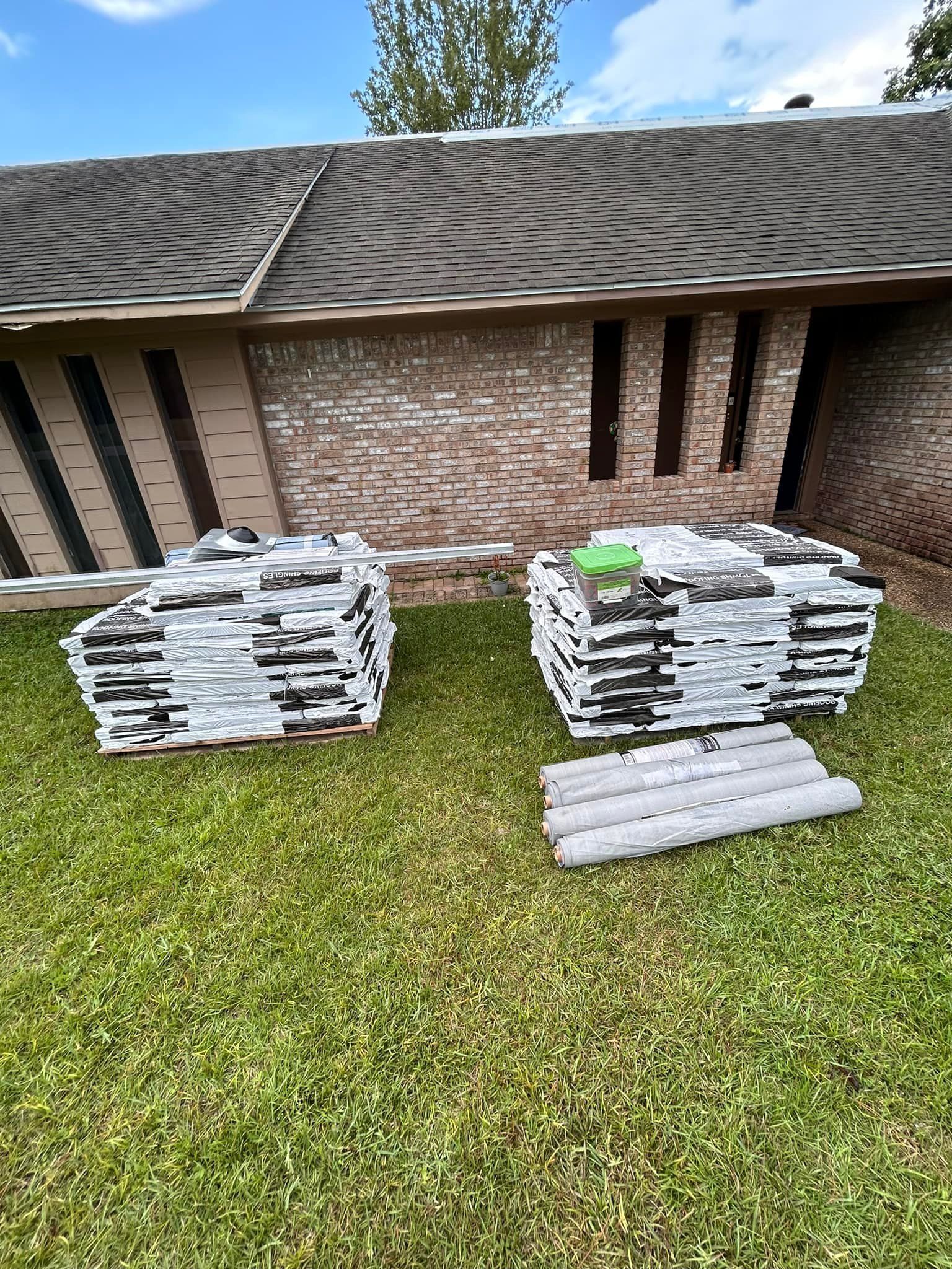 Two stacks of roofing shingles on green grass, beside a brick building with a dark shingled roof.