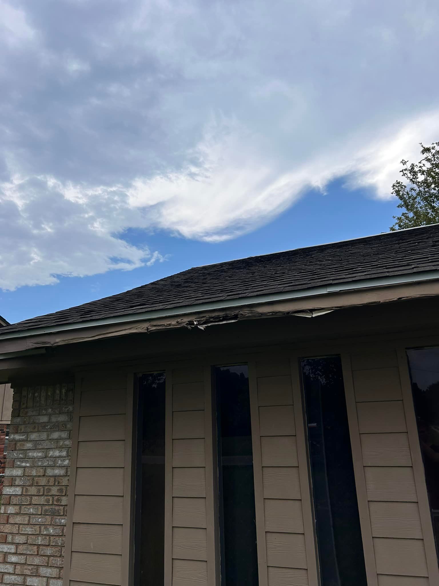Close-up view of a house exterior with dark roof tiles against a cloudy sky. The wall is beige with vertical windows.