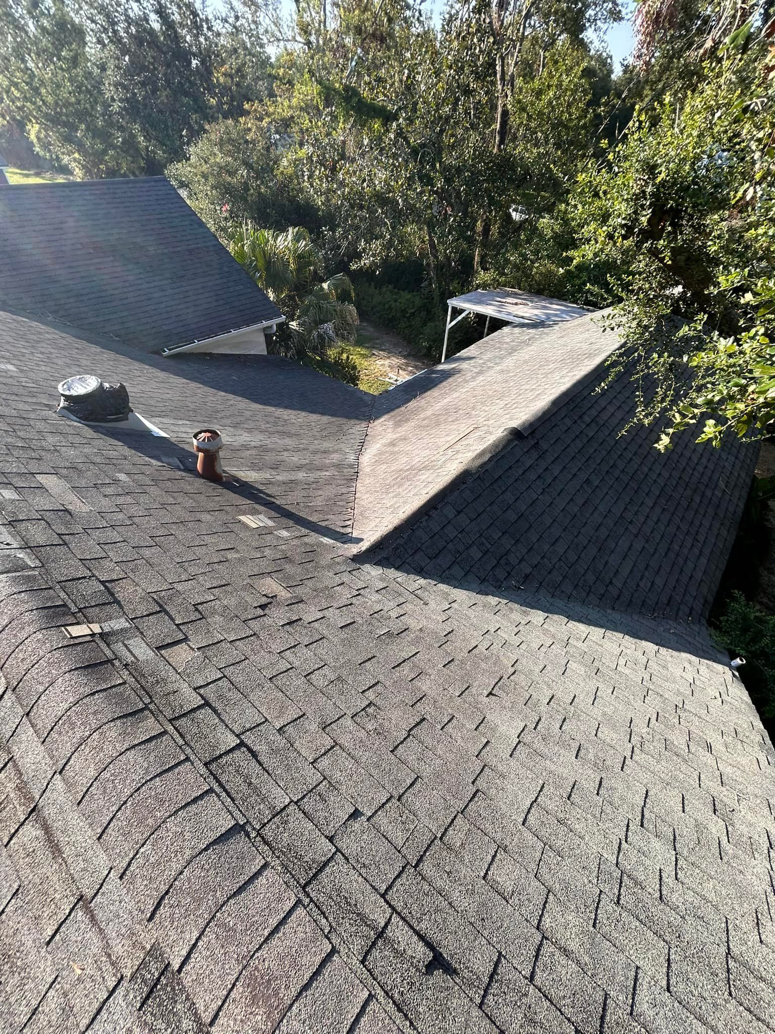 Overhead view of a dark asphalt shingle roof with a chimney, vent pipes, and trees in the background.