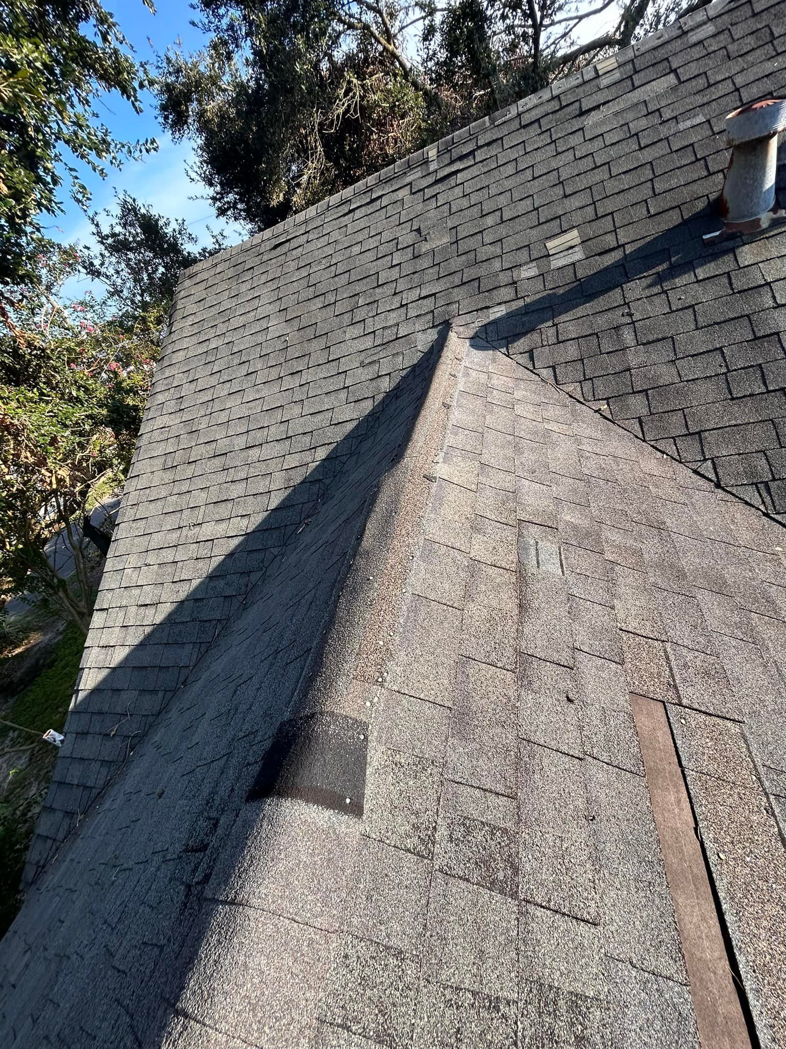 Close-up of a gray asphalt shingle roof with an area of missing shingles and debris, under a blue sky.