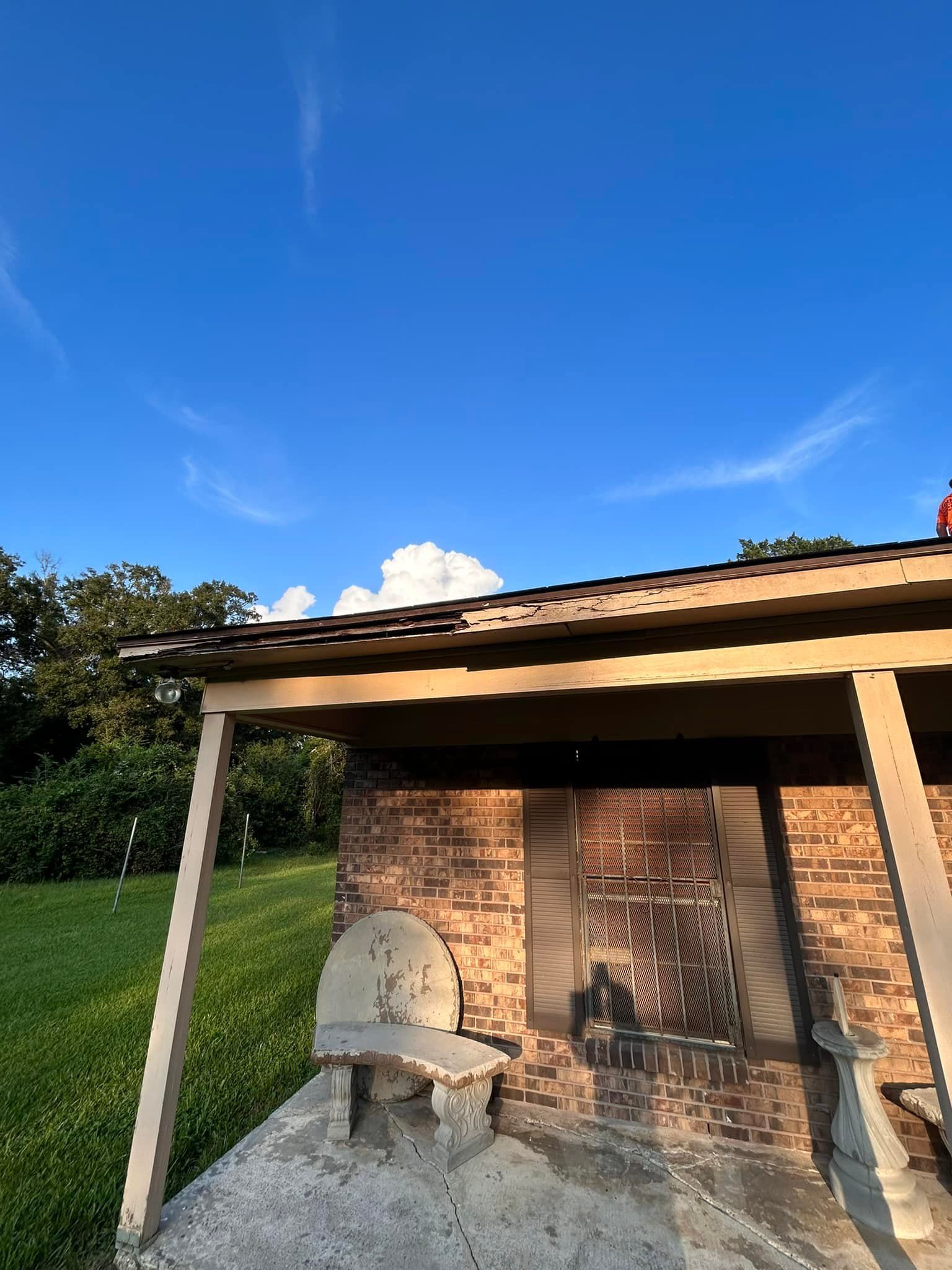 Brick barbeque pit with a roof under a blue sky. A white bench sits in front. Green grass and trees are in the background.
