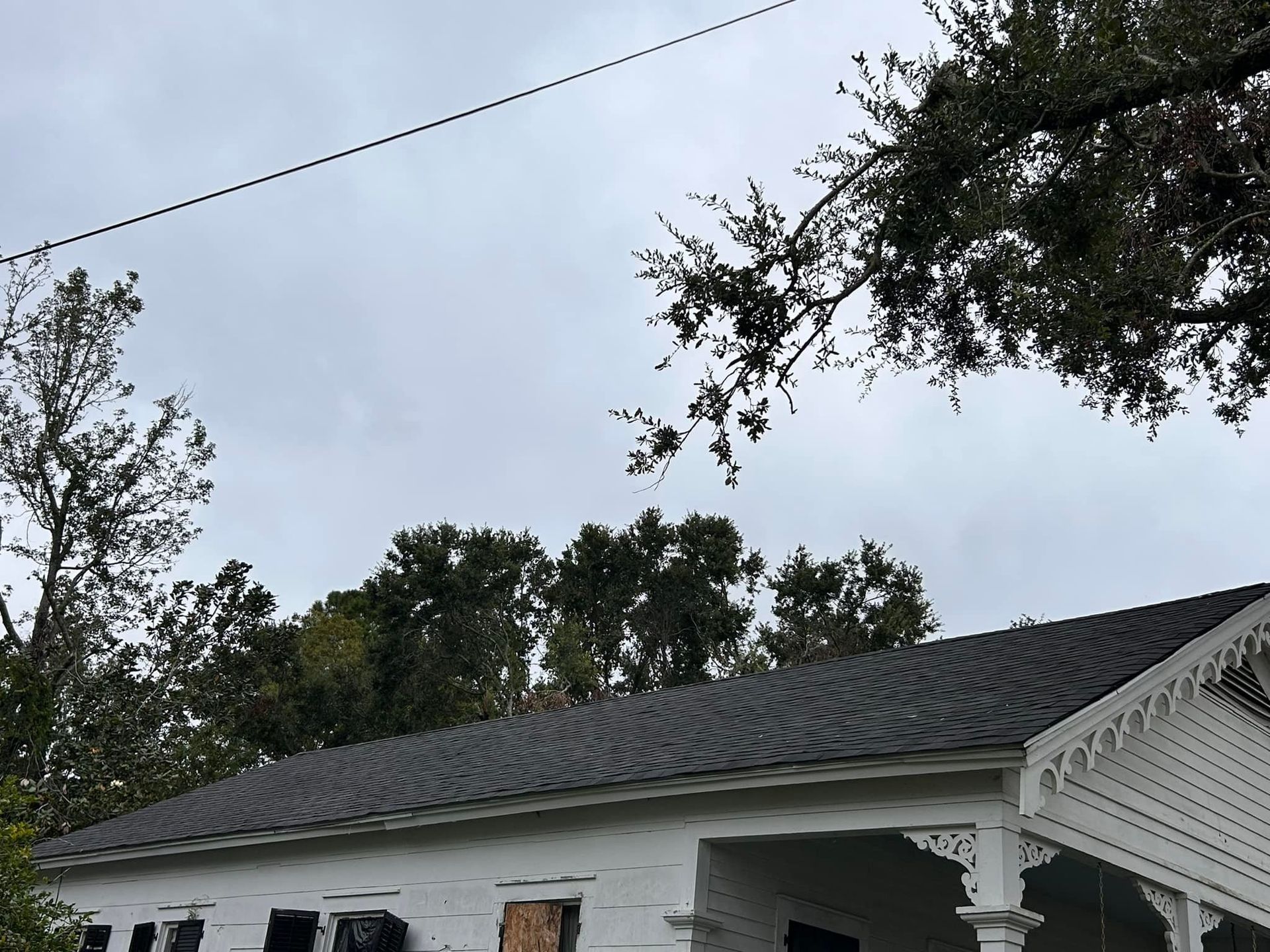 White house with dark shingled roof, boarded windows, and decorative porch. Trees and overcast sky in the background.