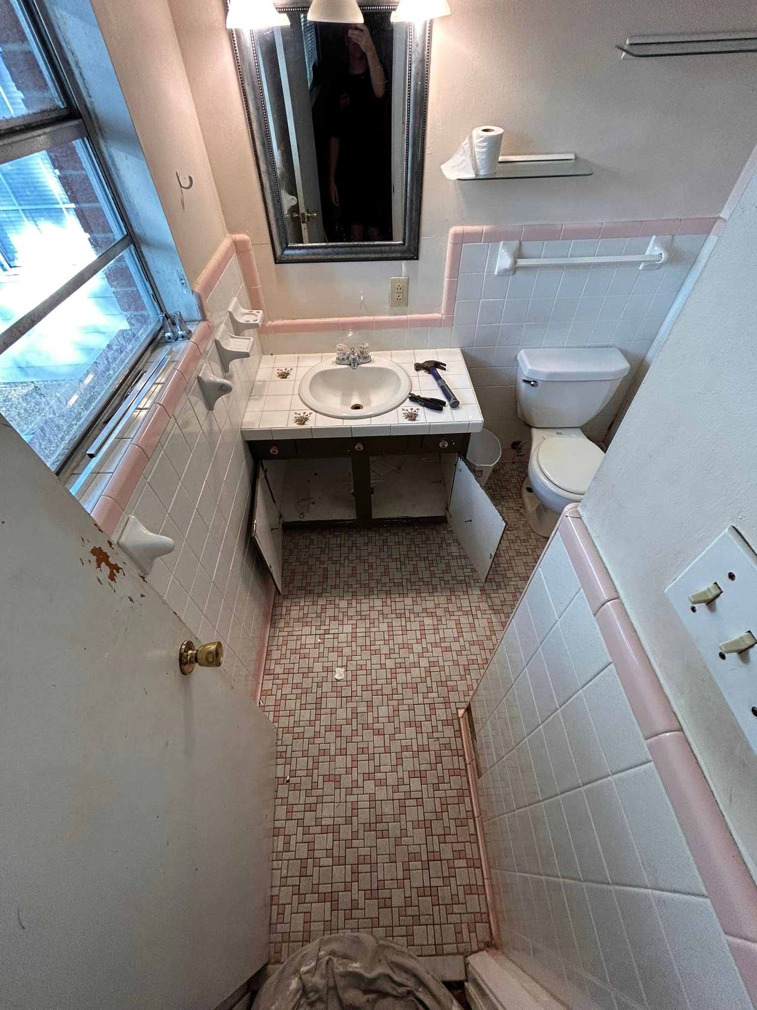 Narrow bathroom with pink and white tile, toilet, sink, and window. The floor tiles are damaged.