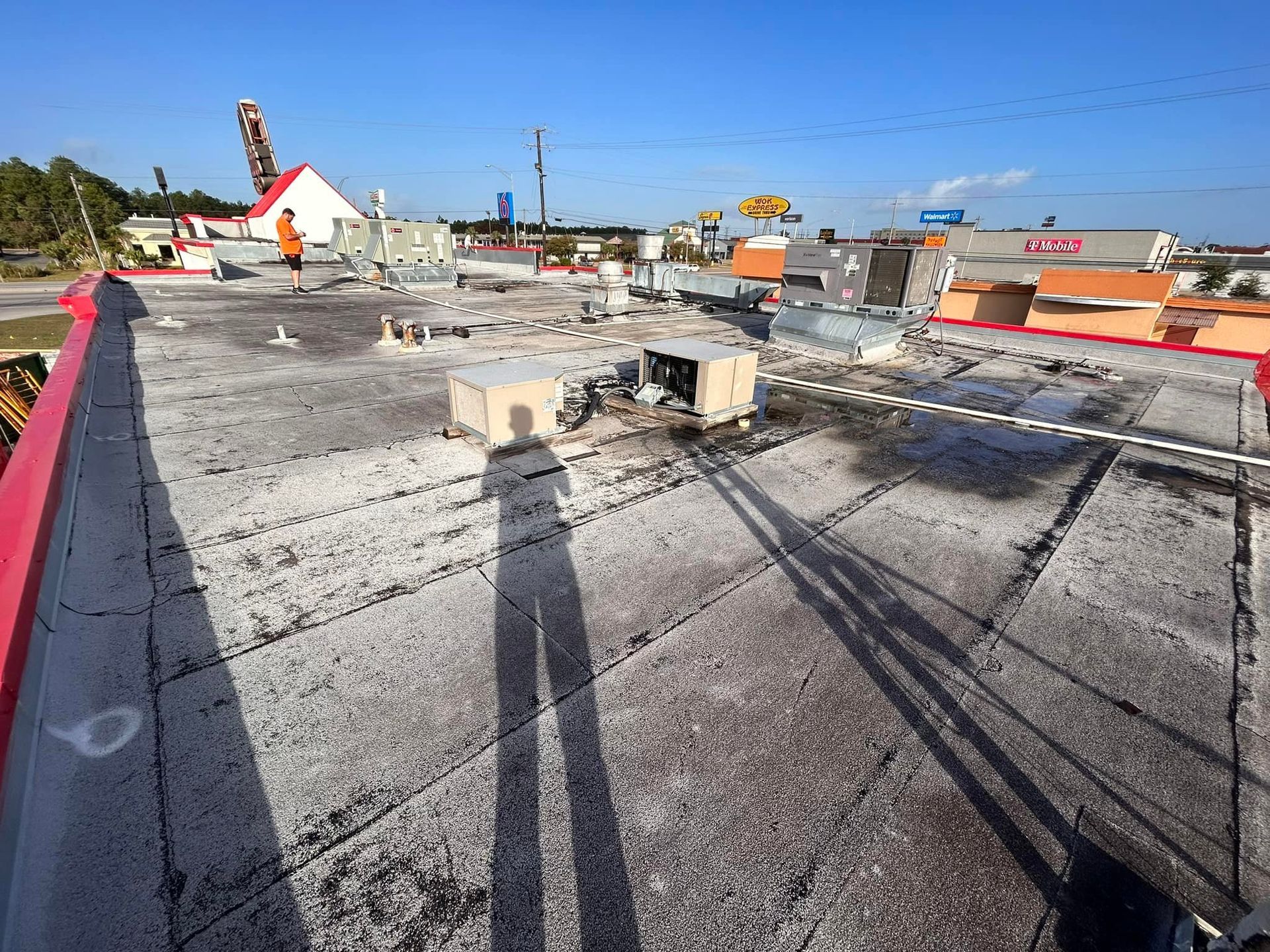 A person's shadow stretches across a weathered flat roof. The roof has various mechanical units, and a cityscape is in the background.