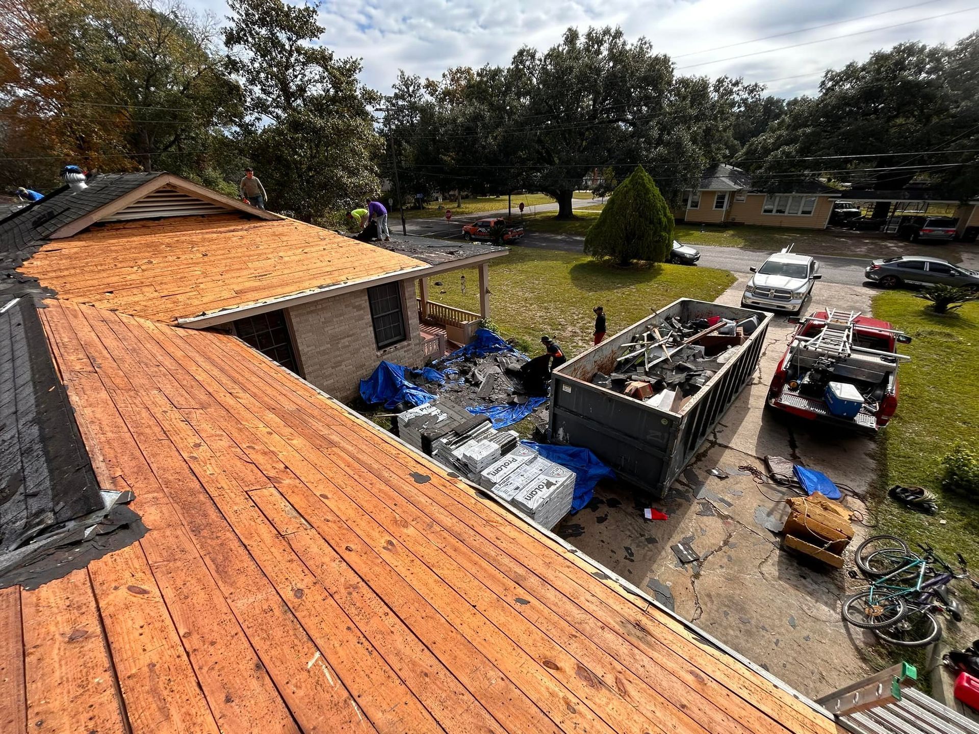Roofing work in progress, debris piled near a house with partially exposed roof.  Workers, a dumpster, and vehicles are visible.