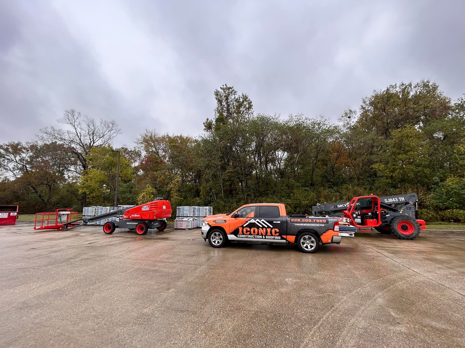 Orange work truck with logo parked near construction equipment on a cloudy day. Trees and bricks are in the background.