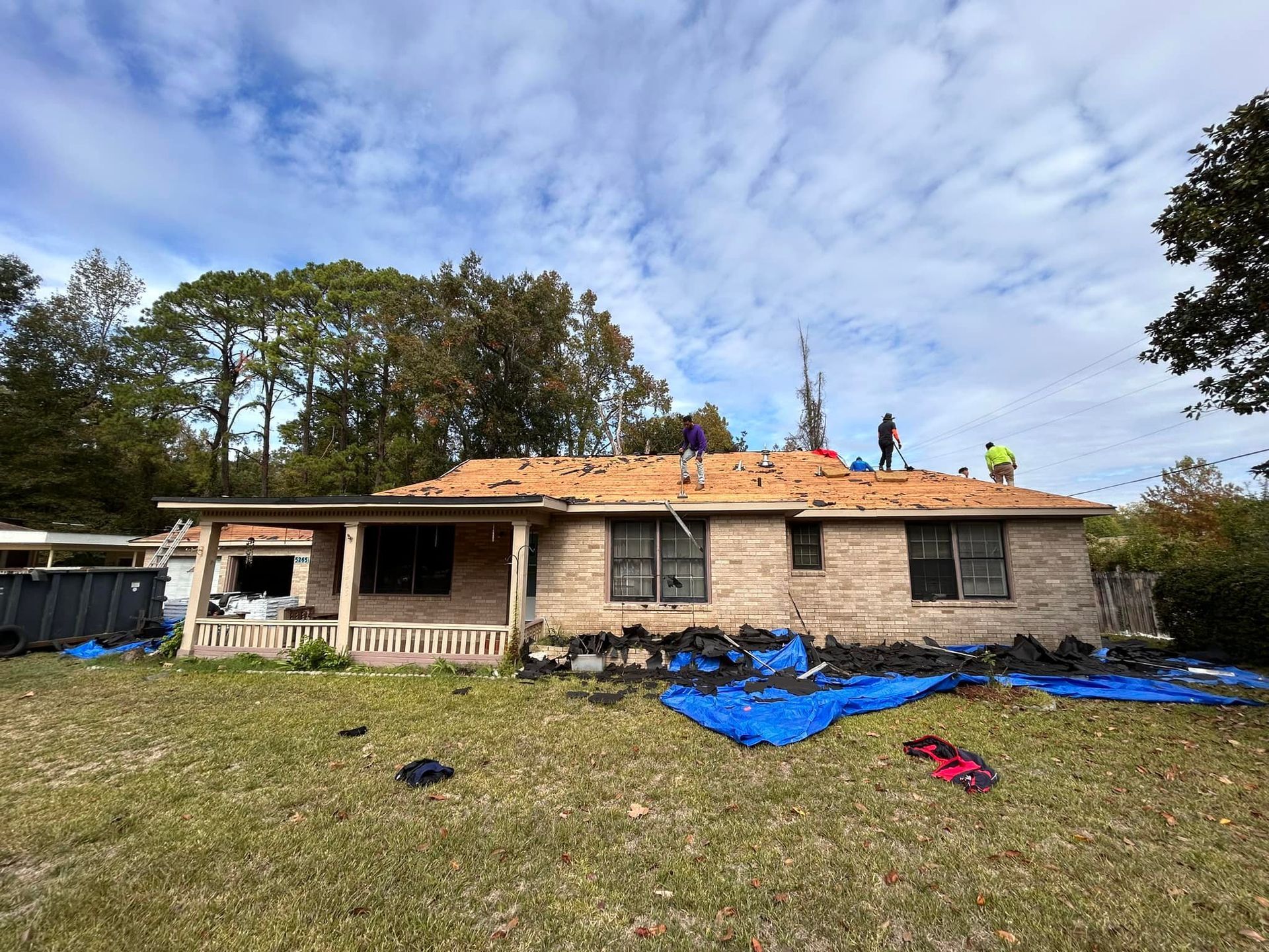 Workers repairing a damaged brick house roof under a partly cloudy sky. Blue tarp covers debris on the lawn.