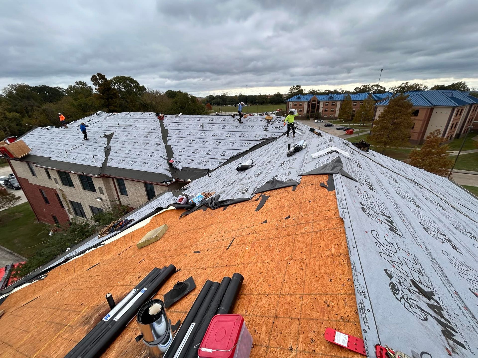 Roofers working on a multi-building complex. The roof is partially covered with new underlayment on an overcast day.