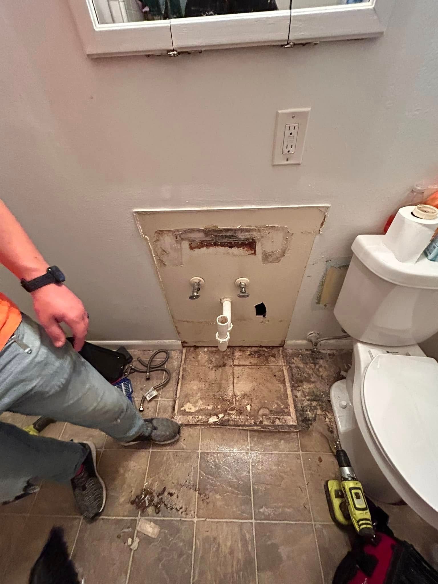 A person removing a bathroom vanity. The partially removed vanity exposes pipes, and the floor has debris. A toilet is visible on the right.