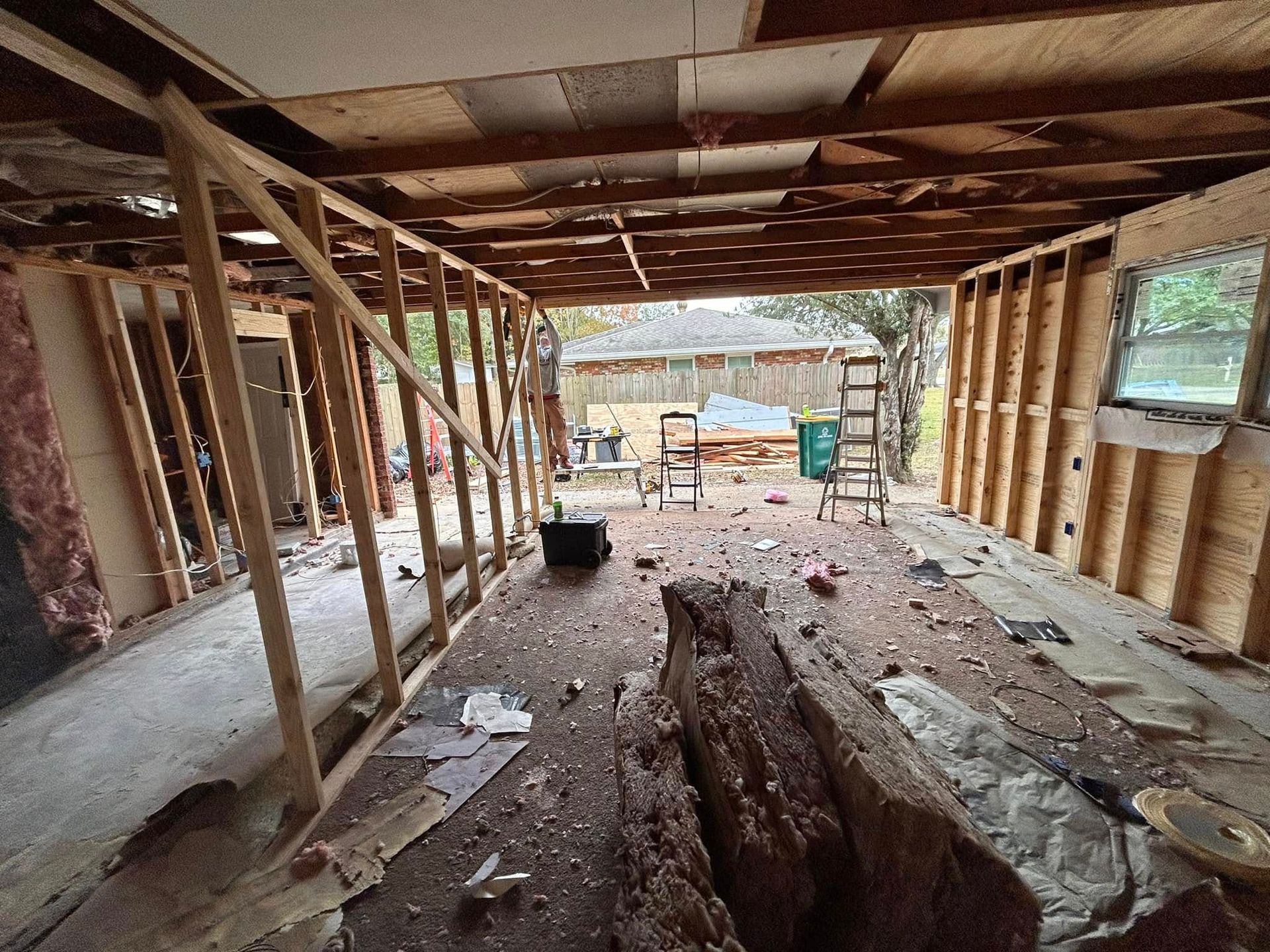Interior view of a house under construction with exposed framing, debris on the floor, and a view of the backyard.